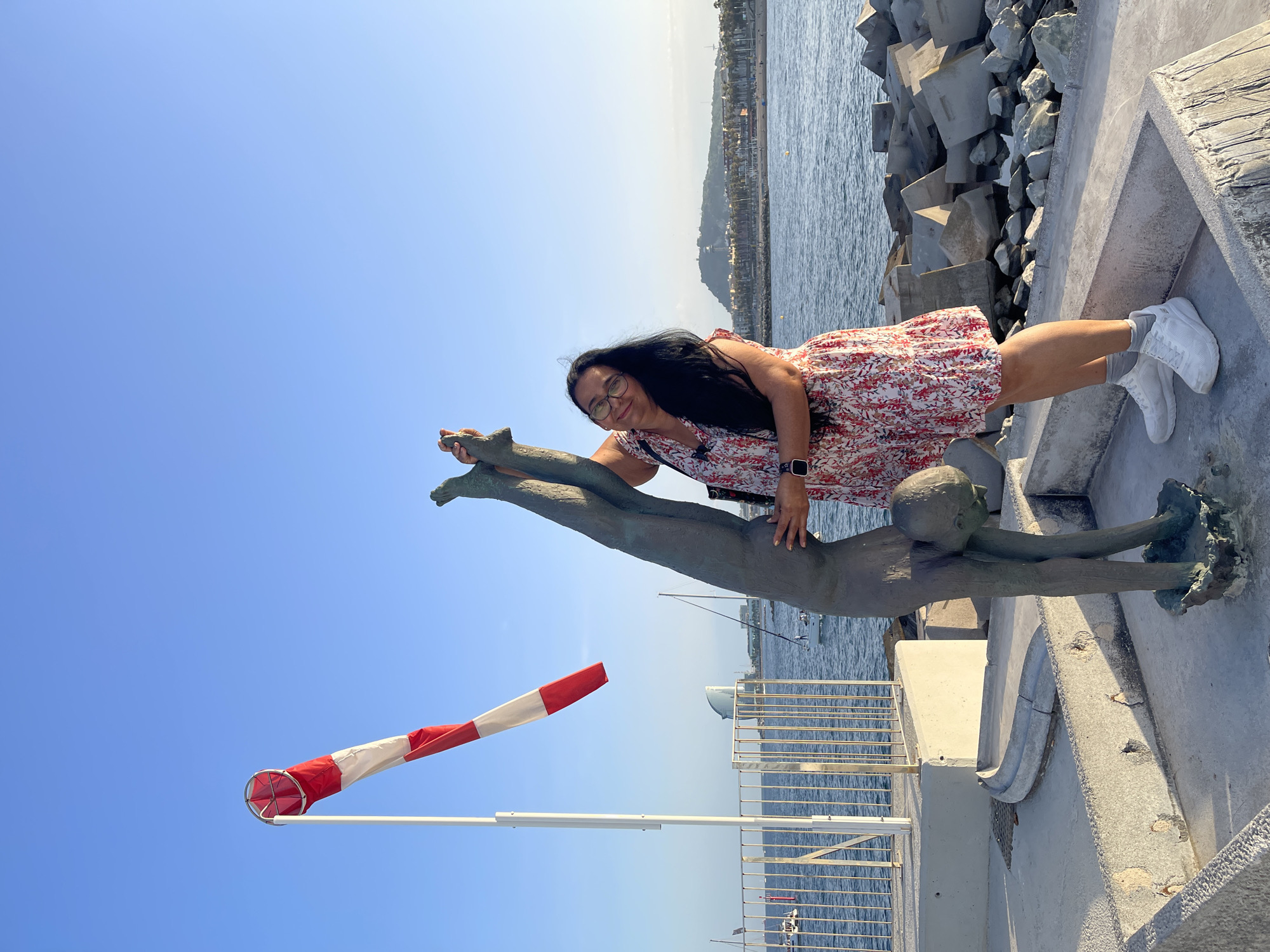 A woman poses with a sculpture of a gymnast in Barcelona, Spain, on a sunny day. The sculpture is of a bronze figure in a handstand pose, with its head bent down. The woman is standing next to the sculpture, with her hand resting on it. She is wearing a floral dress, white sneakers, and glasses. In the background, there is a body of water with boats, a breakwater made of concrete blocks, and a distant city skyline with hills and a tower. A red and white windsock is visible on a pole to the left of the sculpture. The scene appears to be a public promenade or waterfront area.