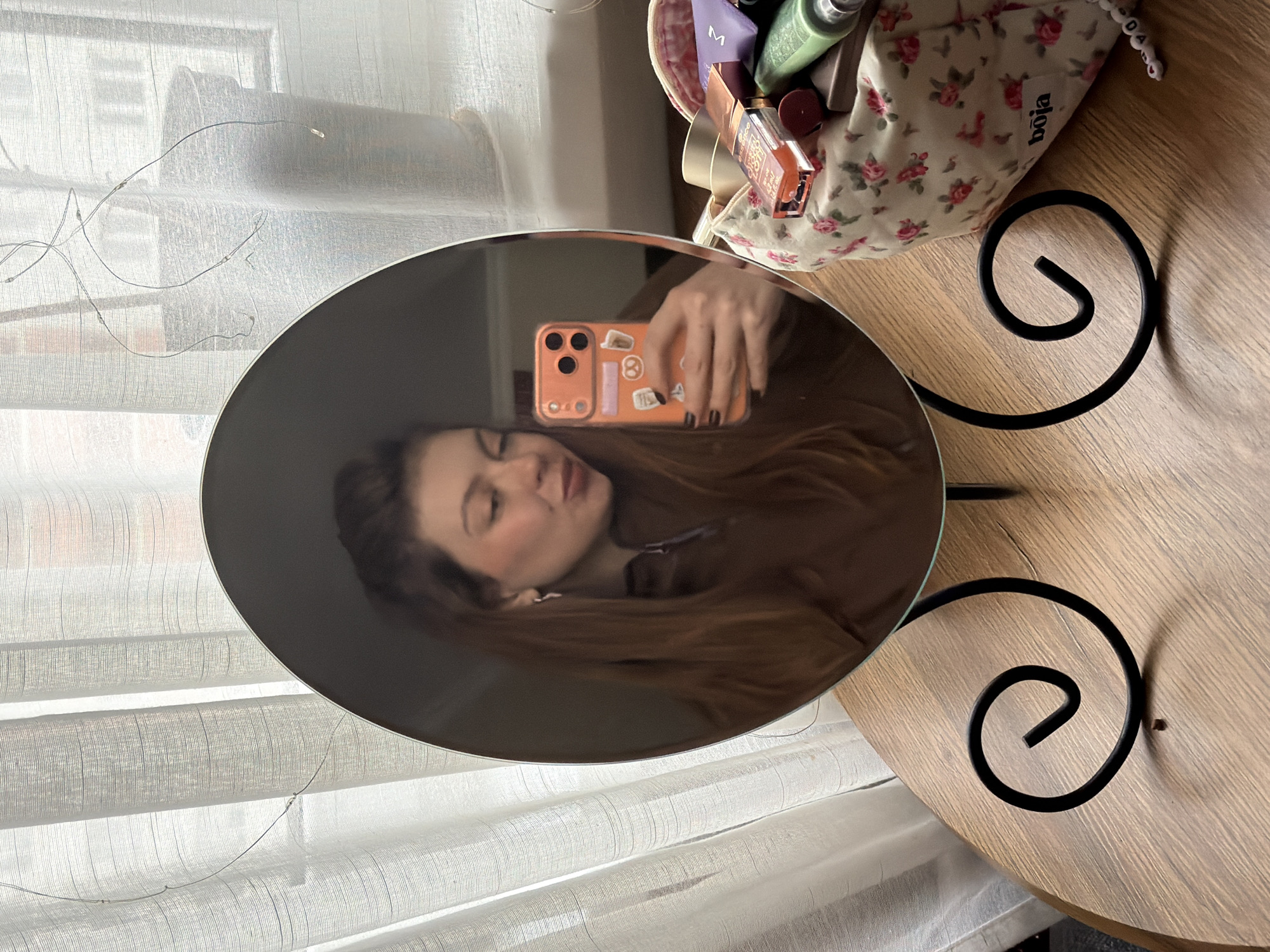 An indoor mirror selfie captures a person with long, dark brown hair and fair skin making a playful "kiss face" expression. The person's hand, adorned with black nail polish, holds an orange smartphone with three camera lenses and several small stickers on its back case. This reflection is seen in an oval-shaped mirror, which rests on a dark, spiral-patterned metal stand atop a light brown wooden table.
To the right of the mirror, a white fabric cosmetic bag with a pink and green rose floral pattern is visible on the table. The text "bōja" is printed on the bag (upside down). The bag is filled with various cosmetics, including a purple tube with a white 'M' logo, a green bottle, and a clear bottle containing orange liquid, possibly from the "ESSENCE" brand, which is partially visible.
In the background, a translucent white curtain with vertical textured lines filters natural light. A string of delicate fairy lights with small bulbs is draped across the curtain. The scene suggests a casual daytime setting, likely within a bedroom or dressing area in Khimki, Russia.