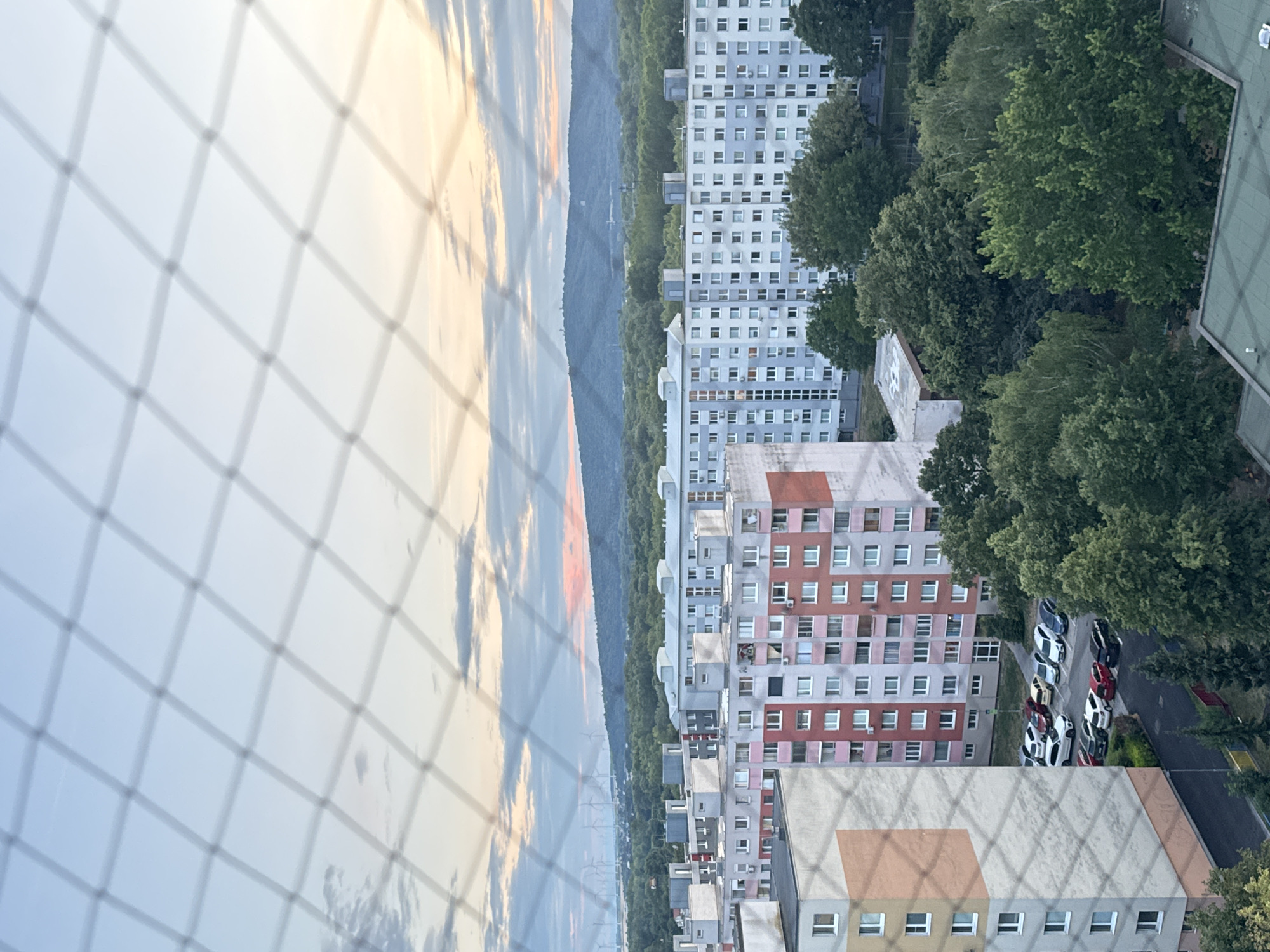 An aerial view of a residential area in Bratislava, Slovakia, captures apartment buildings and a parking lot at dusk. A net is visible in the foreground, obscuring parts of the scene. The sky displays hues of pale blue and pink, with scattered clouds. In the distance, a hilly landscape covered with trees stretches towards the horizon, and a line of wind turbines is discernible. The apartment buildings are characterized by their repetitive window patterns and varied color schemes, with one building featuring a distinctive pink and red facade. Parked cars, including several white sedans and SUVs, are organized in the lot. Lush green trees surround the buildings and dot the landscape.