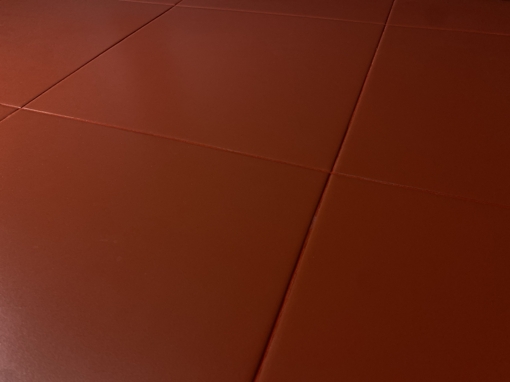 The image is a close-up, abstract view of a textured, reddish-brown surface with visible seams forming geometric patterns. The lighting creates subtle highlights and shadows, emphasizing the texture and the angular divisions of the surface. The image does not contain any discernible elements that would allow for its location to be identified within the city of Perm, Russia, or any other specific geographical location. There are no people, subjects, objects, or actions depicted. The scene is entirely abstract and textural.