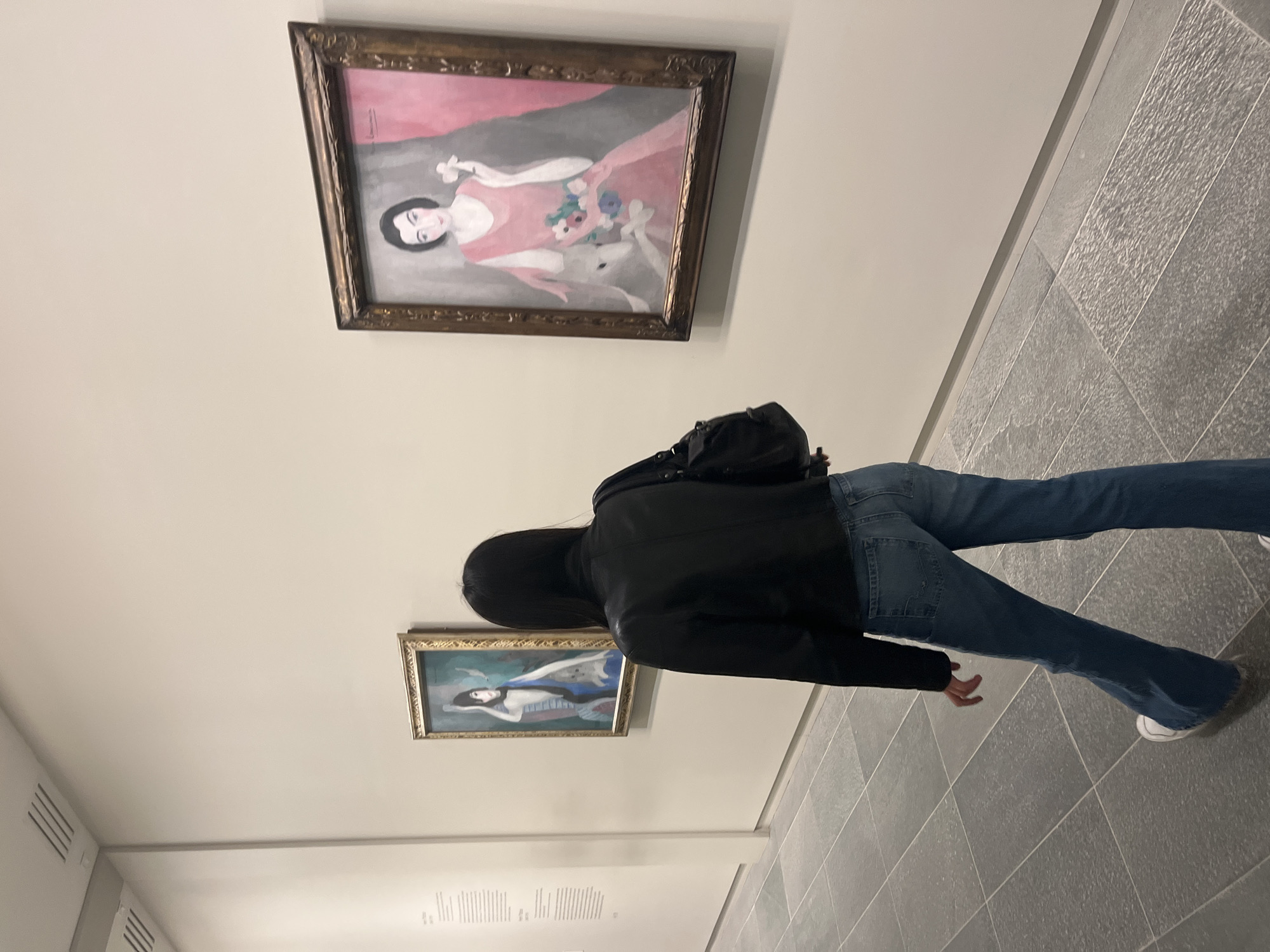 The image presents an indoor scene within an art gallery or museum, likely located in Paris, France. A person, seen from behind, stands centrally, observing two paintings displayed on a light-colored wall. The individual is dressed in a black jacket or hoodie, blue jeans, and white sneakers, with dark hair.
The upper painting, set in an ornate gold-colored frame, depicts a woman with dark hair in a pink or coral-colored dress. She holds a bouquet of flowers, and a light-colored animal, possibly a dog or small deer, is visible at her feet. A signature resembling "Lempicka" is faintly visible in the upper right, suggesting it is a work by Tamara de Lempicka. The lower painting, also in a gold-colored frame, portrays a reclining female figure, possibly nude or semi-nude, with blue and red elements around her.
The gallery space features clean, plain light walls and a textured grey tiled floor. Below the lower painting, there is an area of white wall text, likely an informational plaque, though the specific content is unreadable. The lighting is artificial, consistent with an indoor exhibition space, and the overall atmosphere suggests a quiet moment of art appreciation.