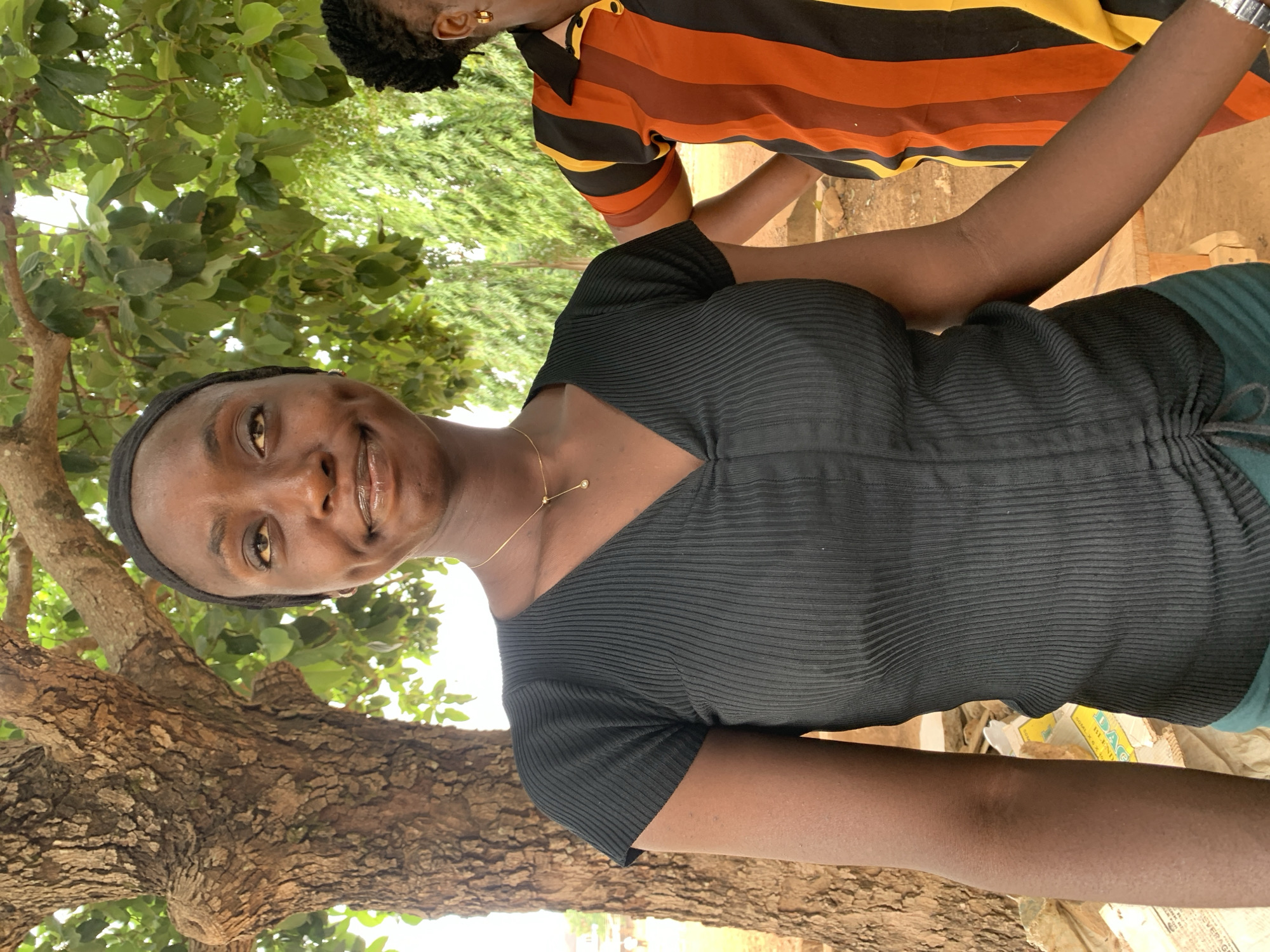 The image shows a dark-skinned woman with a black headband, wearing a black ribbed shirt and a gold necklace. She is smiling and appears to be outdoors, near a tree. Another individual is partially visible in the background, wearing a striped orange, brown, yellow, and black shirt. The location is Maigizo, Nigeria. The ground appears to be slightly dirty. Some discarded cardboard is visible.