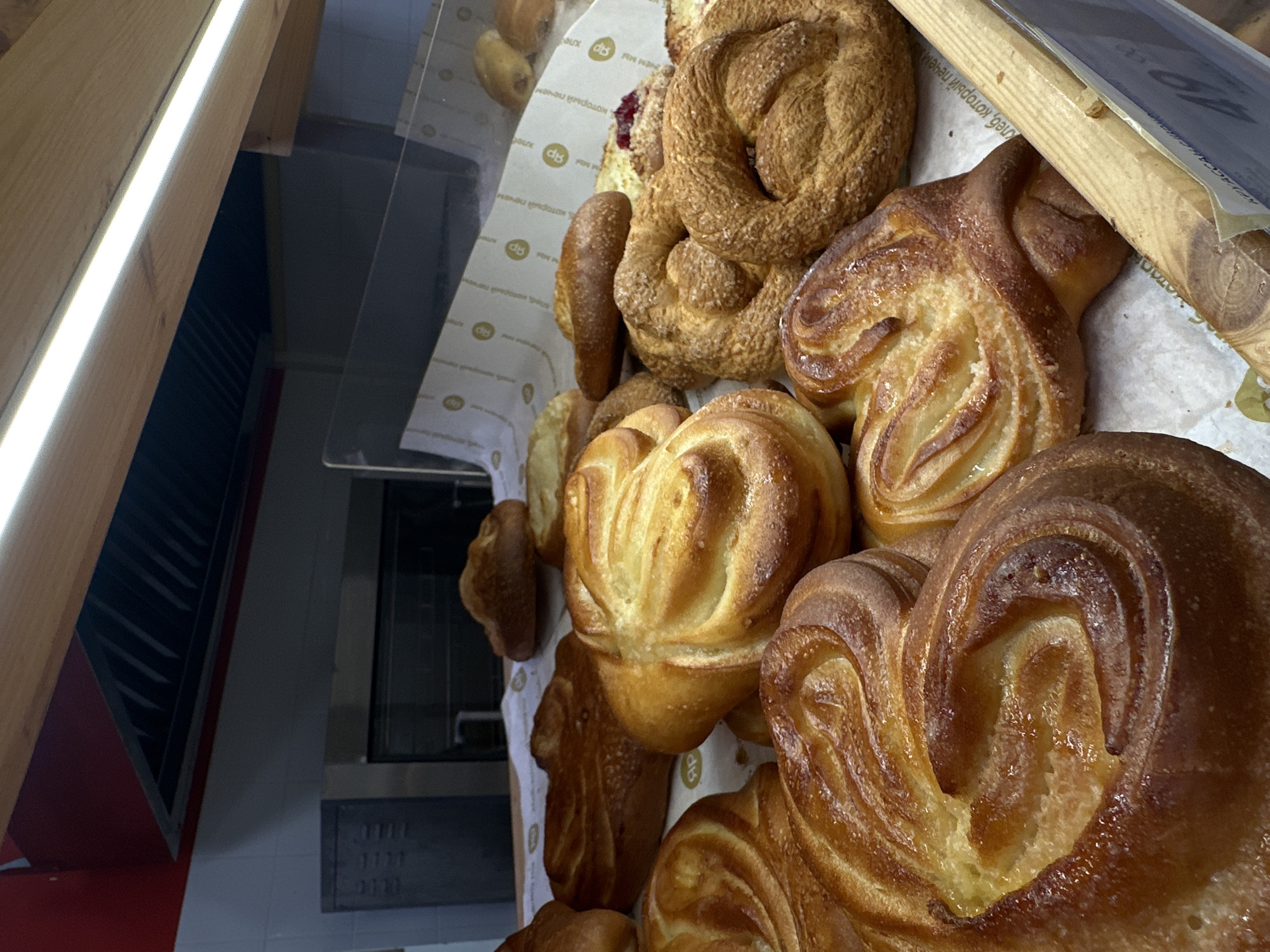 Here is a forensic description of the image:
**Scene:** The image presents a close-up, high-angle view of a display of baked goods, likely within a bakery or café. The pastries are arranged on a white paper lining, possibly in a display case.
**Subjects:** The primary subjects are the pastries themselves, which include a variety of sweet buns and a ring-shaped pastry sprinkled with sugar. The buns display a swirl pattern and are golden-brown in color.
**Objects:**
* Assorted pastries, including buns with a swirl pattern and a ring-shaped pastry.
* White paper lining for the display case.
* A transparent glass display case or shelf.
* A wooden frame or support structure for the display case.
* A price tag partially visible at the top right corner.
**Environment:** The pastries are contained within a transparent display case, indicating a bakery or similar establishment. The presence of kitchen-like equipment, such as oven, along with red panels, suggests it's located in the kitchen or a food retail environment.
**Location:** The text on the paper lining includes the brand name "Яр" and the Russian words "Клеб, который печем". Given the brand name, the location is likely Krasnoyarsk, Russia, where this brand operates.
