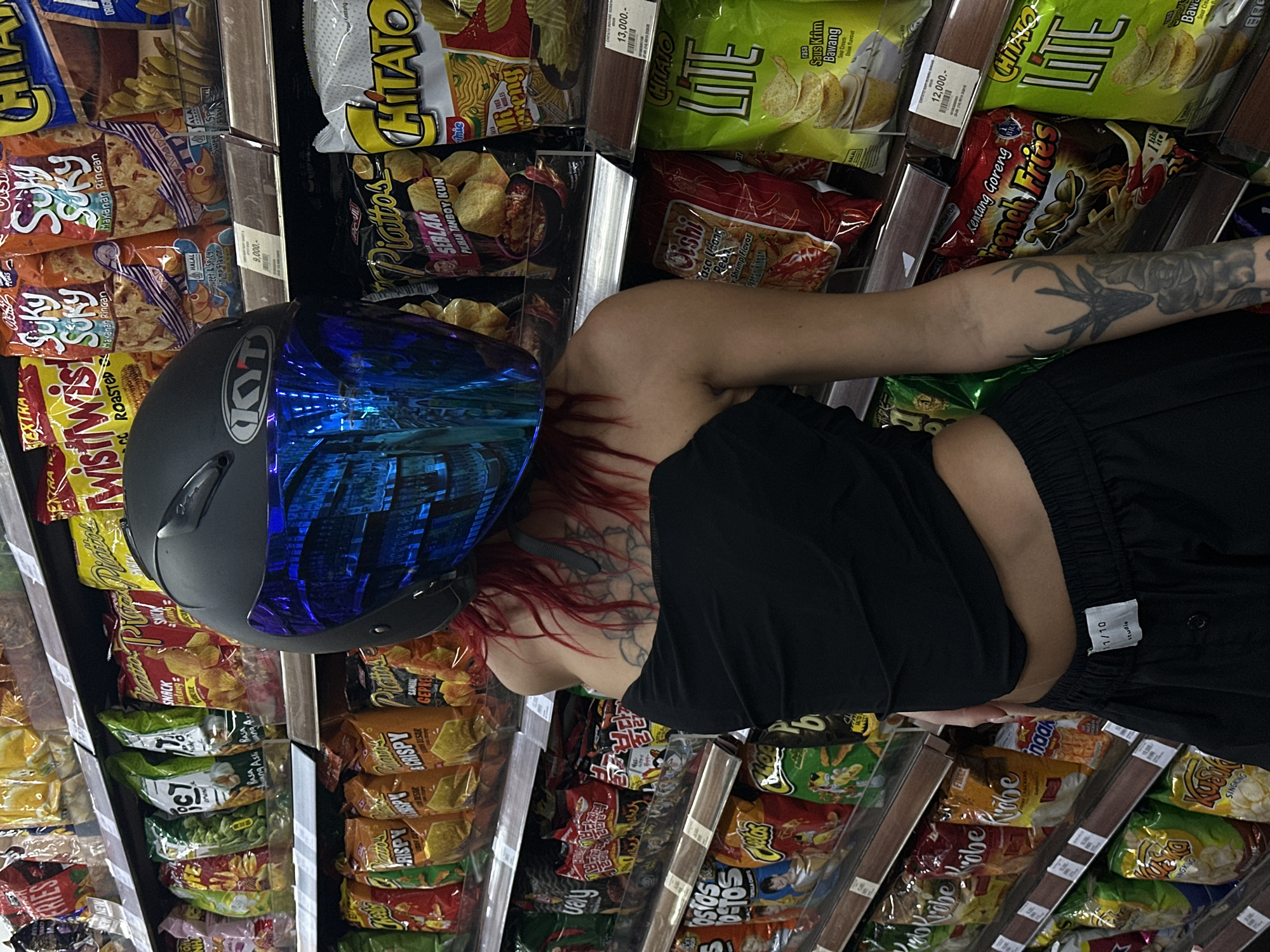 A young woman in a black crop top and black pants stands in front of a grocery store shelf in an aisle filled with various types of snack chips in North Kuta, Indonesia. She is wearing a black and blue motorcycle helmet and has red hair and tattoos on her arms and neck. The shelves are stocked with chips in colorful packaging, including brands like Piattos, Chitato, and Oishi. There are also price tags visible on the shelves.