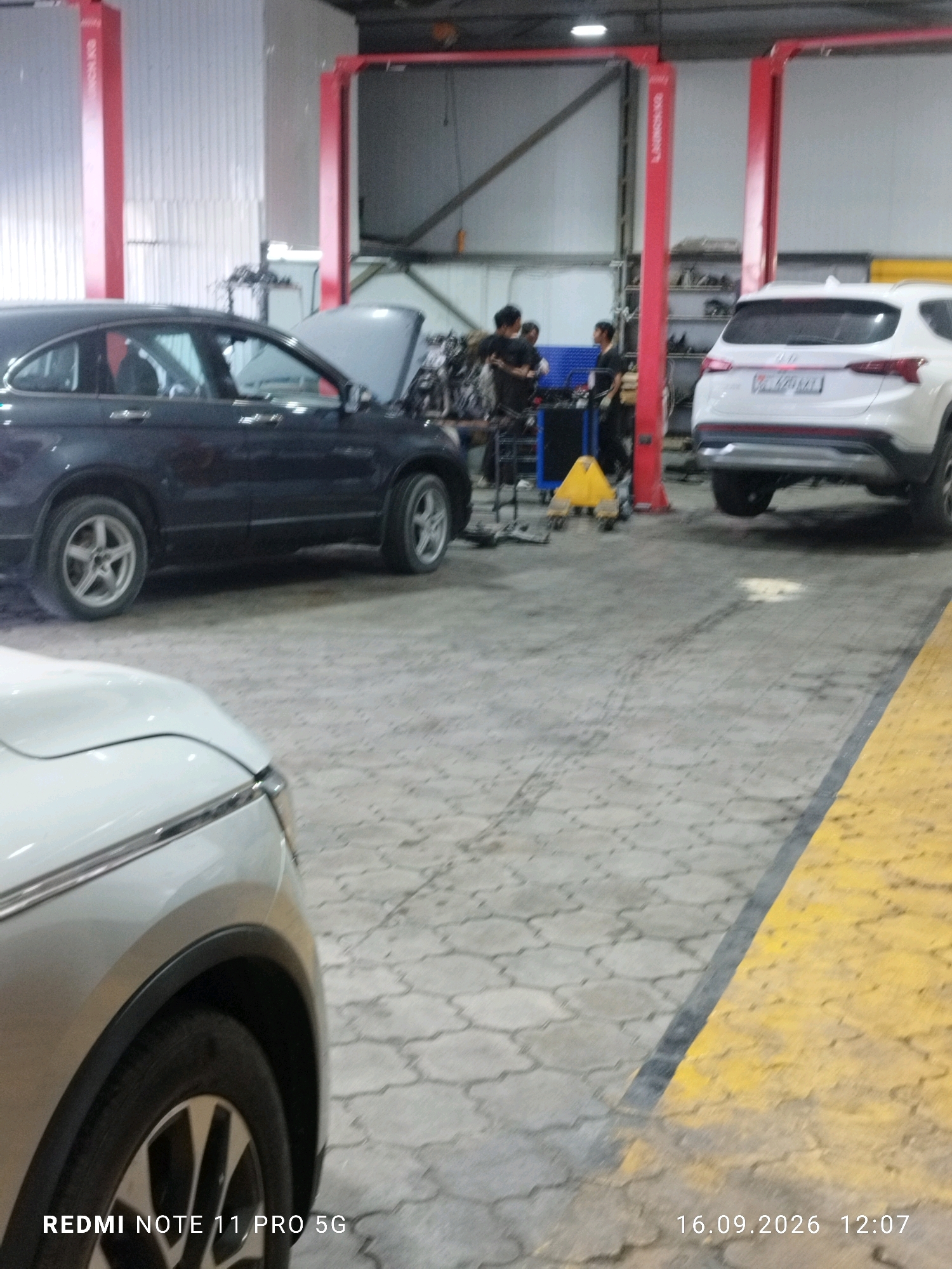 A dark gray SUV is parked in a repair bay with its hood open. The vehicle is on a paved surface that appears to be inside a garage or service center. Two people are visible in the background, appearing to be mechanics working on another vehicle or equipment. A white SUV is also present in the bay to the right. The environment suggests an automotive repair shop.