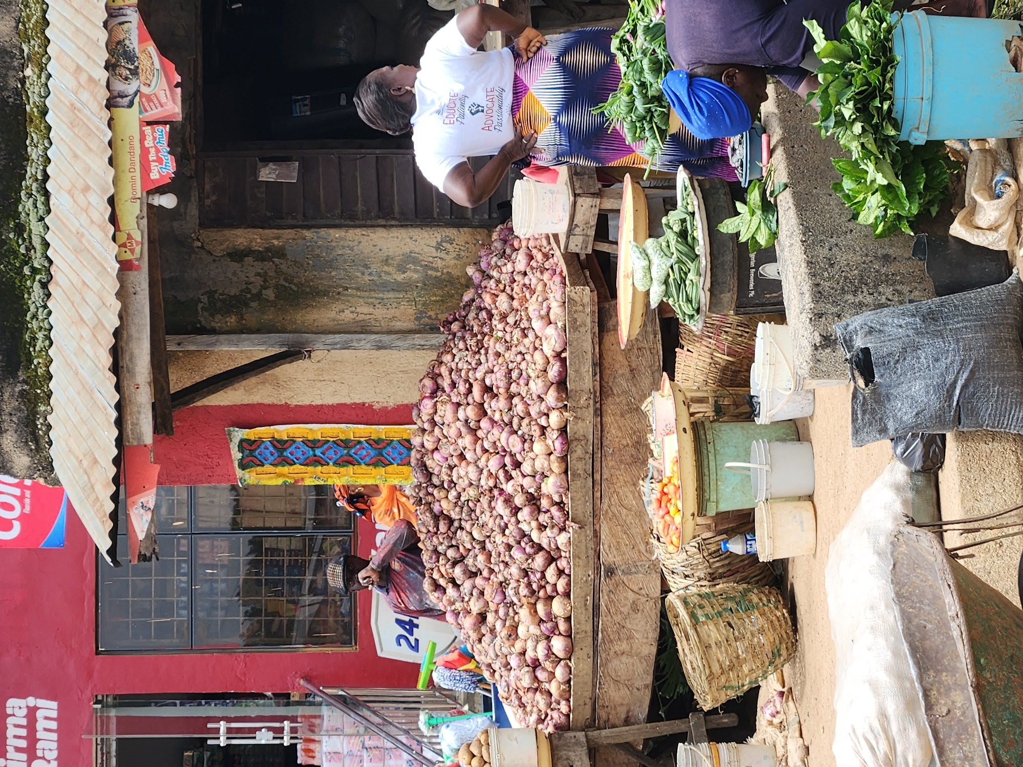 The image depicts an outdoor market scene in Kabong, Nigeria. In the foreground, a large mound of onions is displayed on a wooden stall. To the right of the onions, a woman wearing a white t-shirt and a vibrantly patterned skirt is standing. In the middle ground, various baskets, buckets, and produce, including okra and tomatoes, are visible. A man in a checkered cap is seated behind a stall, and a woman in an orange headscarf is partially visible behind him. In the background, a red building with a window and a corrugated metal roof over a market stall can be seen. The overall environment suggests a bustling local marketplace.