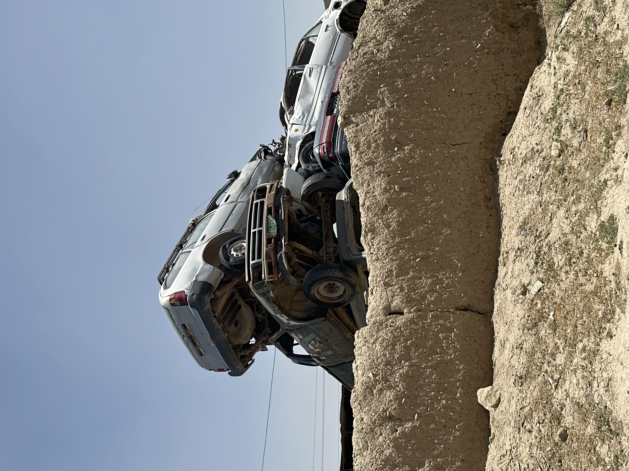 In Kanozai, Pakistan, a junkyard is visible with several rusted and dismantled cars piled high against a clear blue sky. The cars are stacked precariously on top of each other, with some appearing to be station wagons and others sedans. The junkyard is located behind a large, rough-textured wall made of clay or mud. The ground in front of the wall is dry and dusty, with sparse vegetation. Power lines are visible in the background. The scene suggests a location where old or damaged vehicles are brought for scrap or disposal.