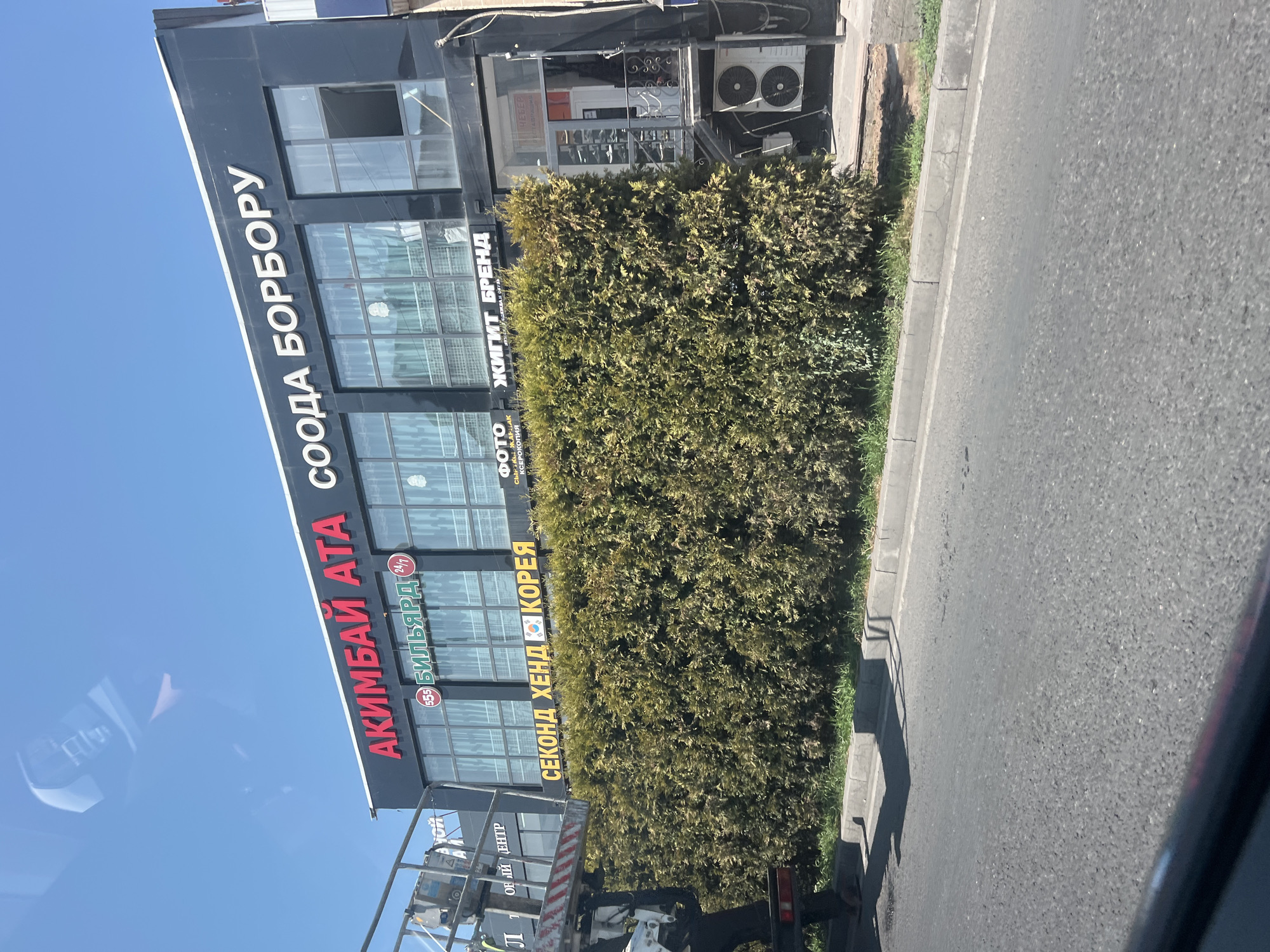 The image shows a street view in Bishkek City, Kyrgyzstan, featuring a two-story building with a black facade. The upper floor has large windows and signage in red and white. Prominent signs include "АКИМБАЙ АТА" in red and "СООДА БОРБОРУ" in white. Below these, smaller signs are visible, one in red reading "(555) БИЛЬЯРД 24/7" and another in yellow reading "СЕКОНД ХЕНД KOREA" with a South Korean flag. The lower part of the building has a glass storefront with railings and an air conditioning unit mounted on the exterior wall. A dense green hedge runs along the front of the building, bordering the street. The street is paved and appears to be a typical urban road. A street light or utility pole is partially visible on the left side, and a portion of a vehicle or construction equipment is also present on the far left. The sky is clear and blue, indicating daylight.