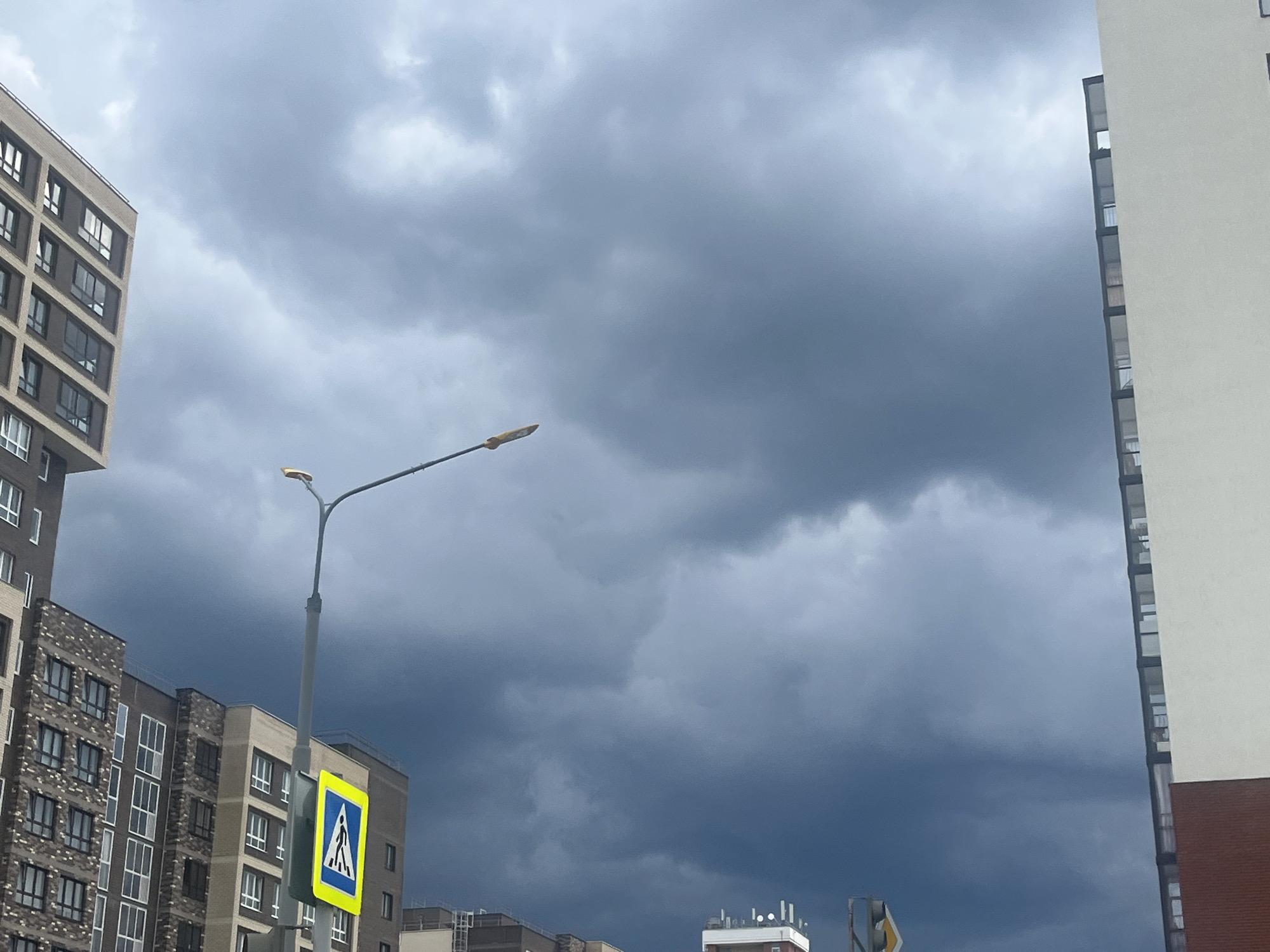 The image shows a street scene in Yekaterinburg, Russia, under a dark and cloudy sky, suggesting an impending storm. In the foreground, a pedestrian crossing sign is visible, attached to a street lamp. To the left, a modern apartment building with many windows is partially visible. On the right, another tall building with a white facade dominates the scene. The overall impression is one of urban infrastructure set against a dramatic weather backdrop.