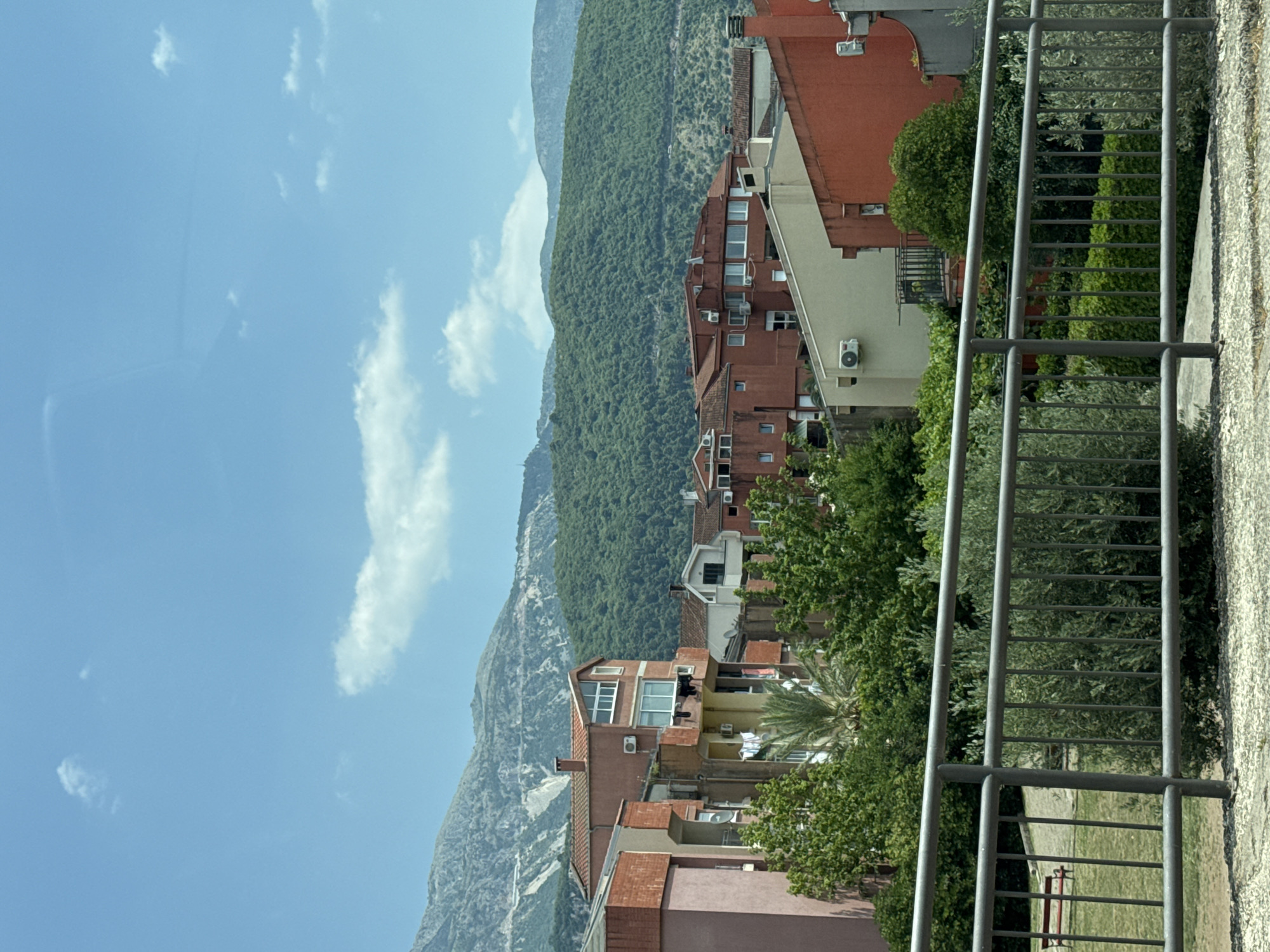 In Budva, Montenegro, a residential area features buildings with terracotta roofs and orange, pink, and beige facades. The structures, some with visible air conditioning units and clotheslines, are nestled against a backdrop of lush green, tree-covered hills. Further in the distance, a light-colored, rugged mountain range dominates the horizon under a bright blue sky dotted with scattered white clouds. A dark metal railing runs across the foreground, separating a paved area from a green space with trees and a red park bench.