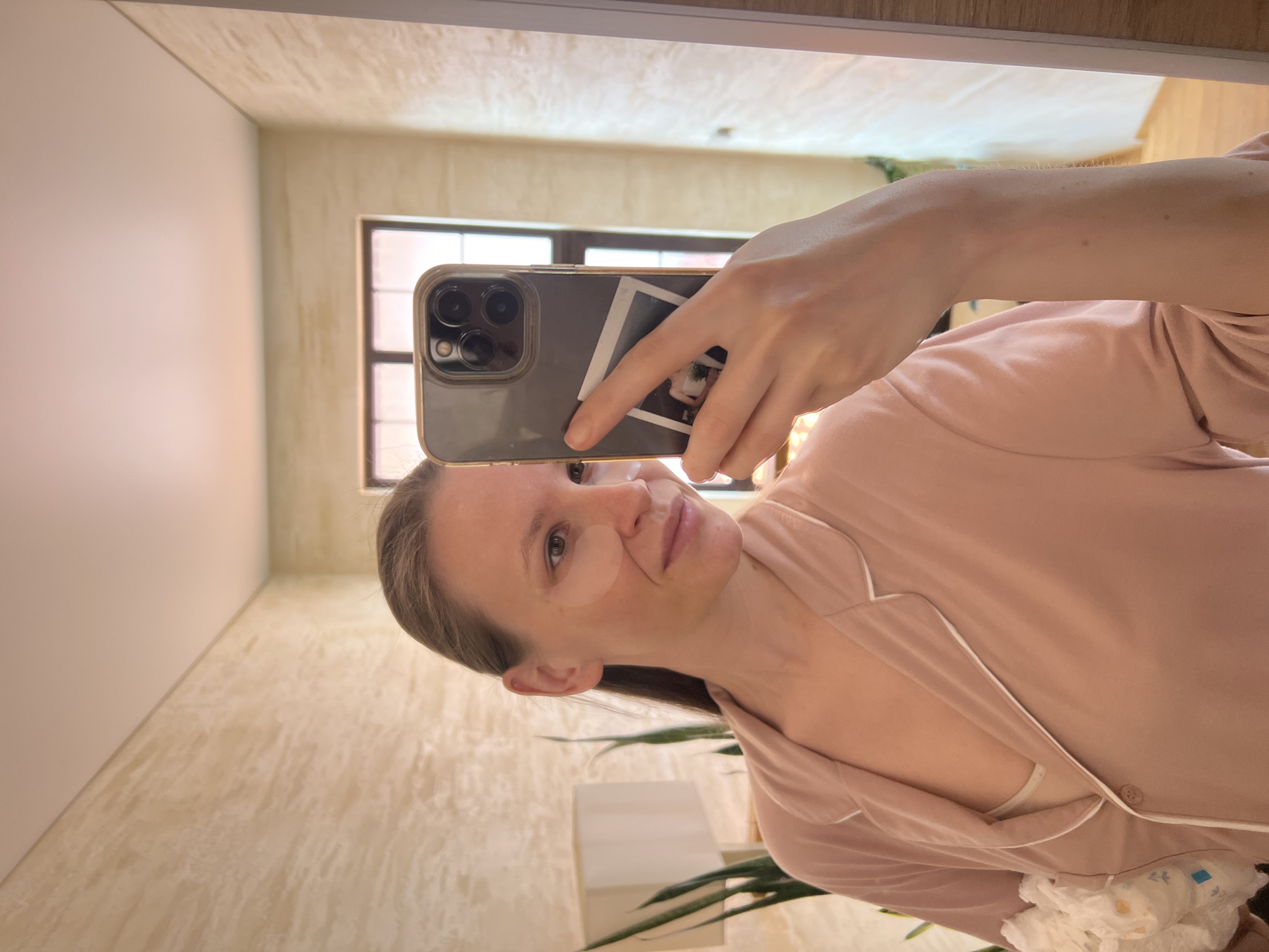 A woman is taking a selfie in a mirror. She is wearing a pink pajama set and has eye patches on her face. She is holding a smartphone with a photograph taped to the back. The room appears to be a bathroom, with textured walls and a window. There is a houseplant visible in the reflection. The specific location in Gelendzhik, Russia cannot be determined from the image.