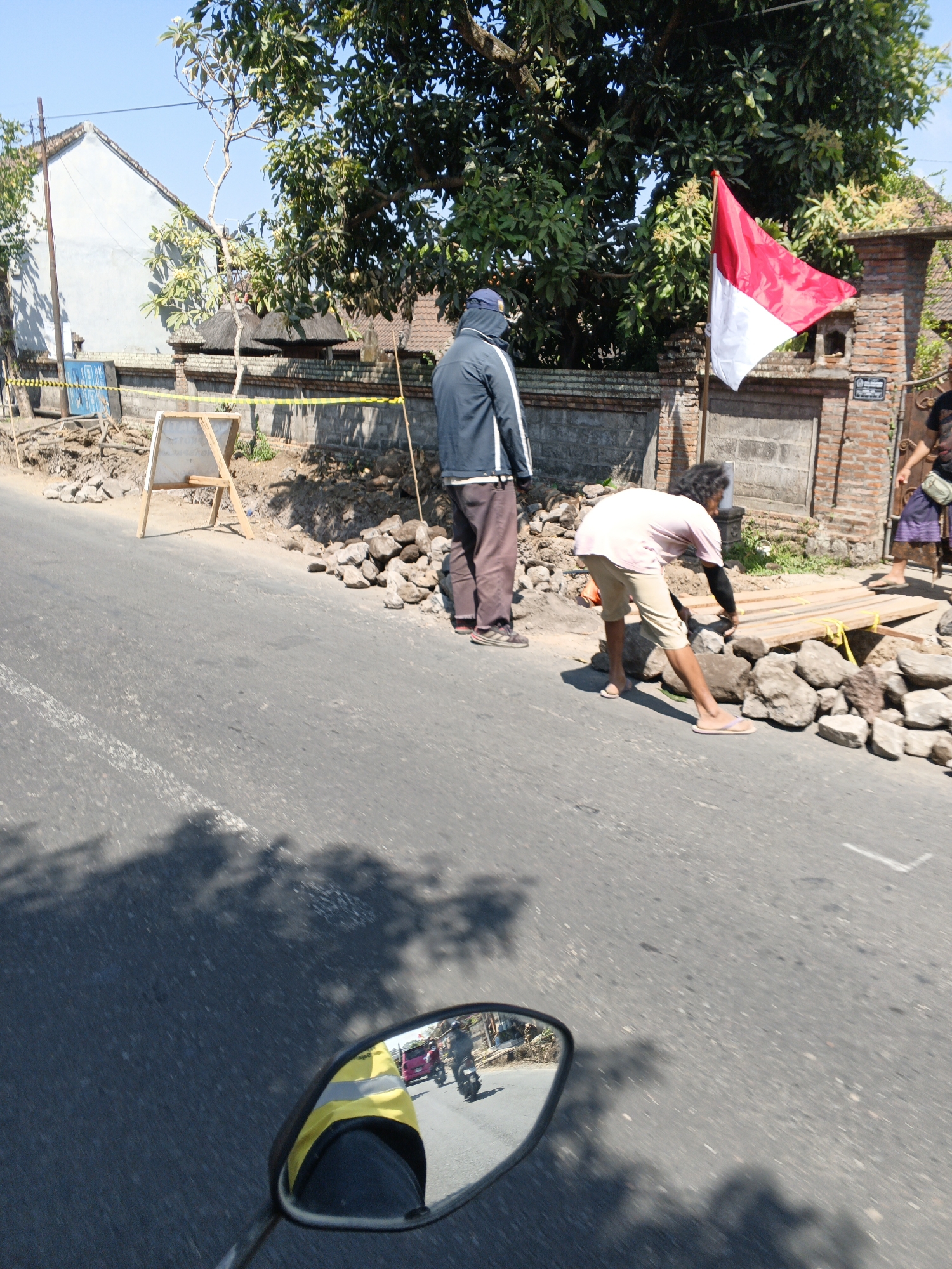 The image captures a scene on a paved road in Sukawati, Indonesia, possibly near a construction or repair site. The road is flanked by a low brick wall and vegetation. A person in the foreground, wearing a light pink shirt and tan shorts, is bent over, appearing to adjust or place wooden planks. They are barefoot with sandals on the ground. To their left, another person stands facing away, dressed in a dark blue jacket with white stripes and dark pants. A red and white Indonesian flag is prominently displayed on a pole to the right of the first person. In the background, a white building with a tiled roof and some trees are visible. The road shows signs of disturbance, with piles of rocks and construction barriers. The reflection in a motorcycle's rearview mirror, in the bottom right of the frame, shows another motorcycle and a pink vehicle, indicating ongoing traffic. The sky is clear and blue.