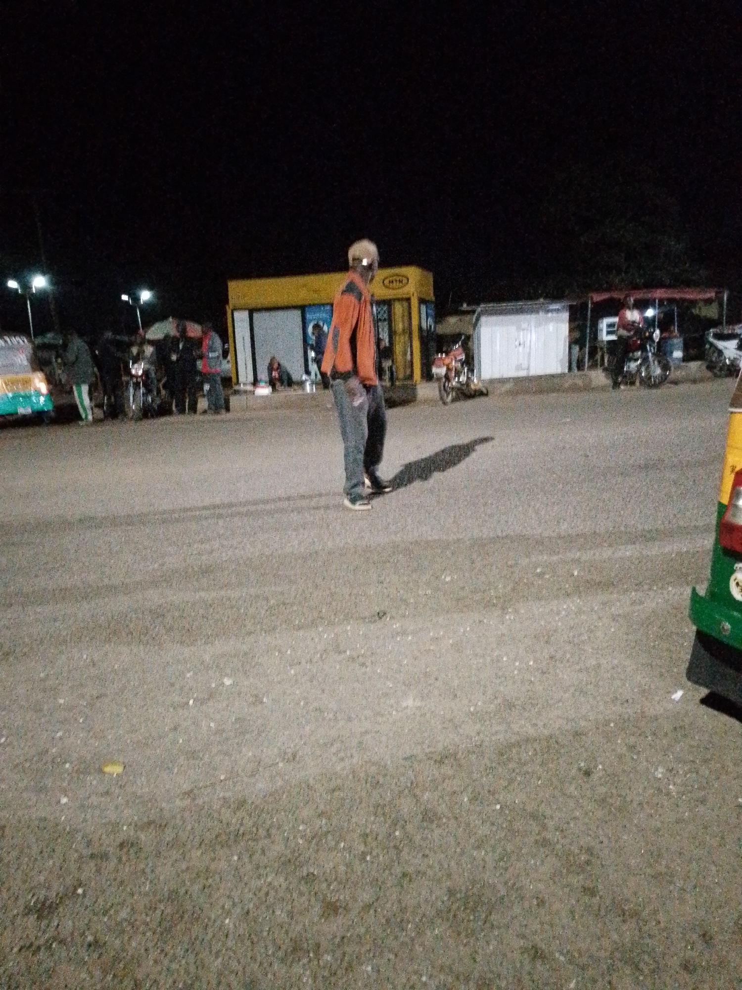 In the city of Bukuru, Nigeria, at nighttime, a man wearing an orange jacket and dark pants walks across an asphalt road. The man's face is obscured by shadow and his stance is slightly hunched. He is casting a long shadow to his left. Behind him, a bright yellow kiosk with the letters "MTN" is visible. Several motorcycles are parked in front of the kiosk and to the right. Further back, a white structure with a red-tarped awning is present, and more motorcycles are parked in front of it, with people standing around them. A green and yellow vehicle, possibly an auto-rickshaw, is partially visible in the lower right corner of the frame. The scene is illuminated by artificial lighting, creating a dimly lit atmosphere.