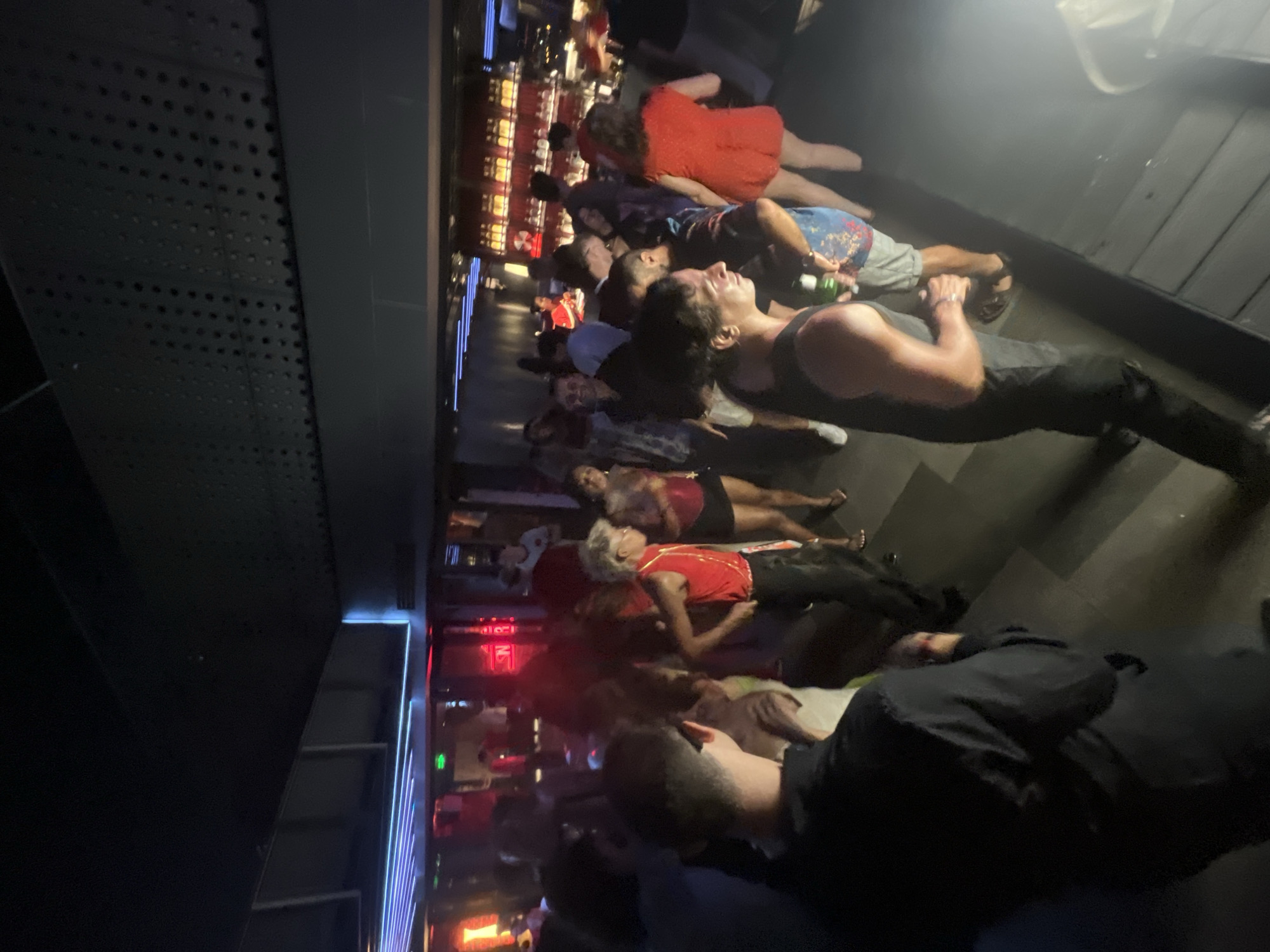 The media file displays a group of people lying down on a dark, glossy floor, likely indoors. The scene is dimly lit, with some areas illuminated by vibrant red and blue neon lights, suggesting a bar, club, or entertainment venue. The people are casually dressed, with some wearing tank tops, shorts, and t-shirts. One person is holding a green bottle. The overall atmosphere appears relaxed, possibly a break from an event or activity. The orientation of the image is rotated, with the subjects appearing to lie down from top to bottom. The setting shows a reflective floor and a textured dark wall or structure on the left. The lighting suggests it is nighttime. There are no visible signs with text in this frame. The location is identified as Đà Nẵng, Vietnam.
