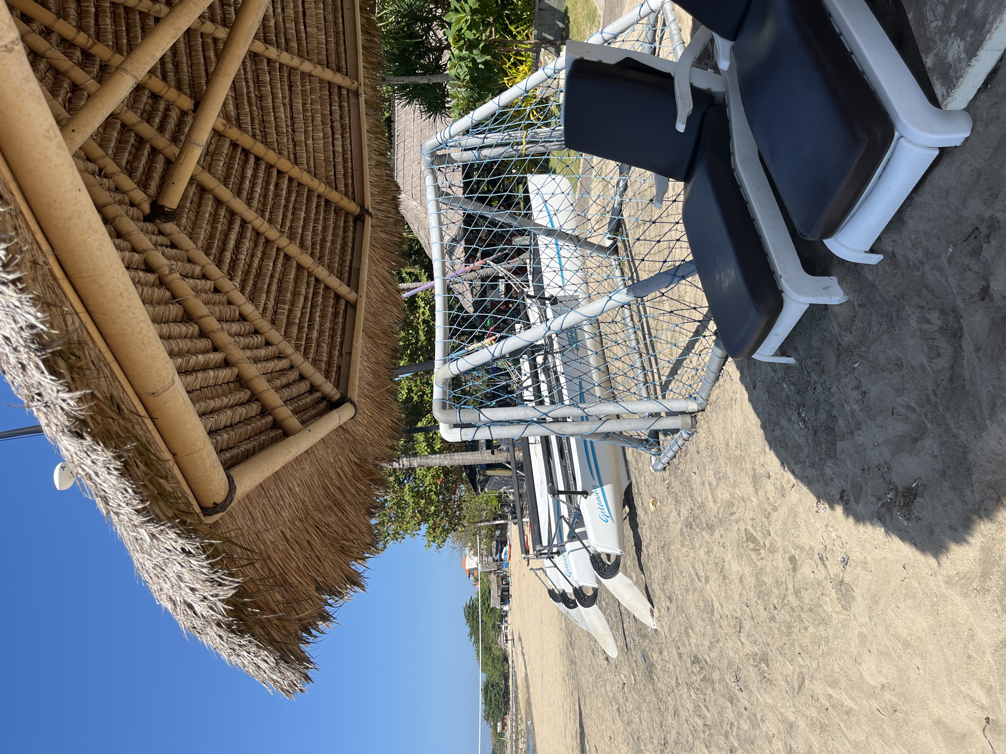 This image depicts a beach scene in Kuta Selatan, Indonesia, under a bright, clear blue sky. In the foreground, two dark-cushioned lounge chairs are positioned on the sand, facing away from the viewer. Behind them, a portable soccer goal with a blue net is set up. To the left of the goal, three white kayaks are lined up, their hulls resting on the sand. Each kayak has the word "Getaway" printed on its side in blue lettering.
A thatched-roof shelter with visible bamboo supports provides shade for the lounge chairs and the area around the soccer goal. The beach itself is sandy with some scattered debris. In the background, beyond the immediate beach area, there are trees and some buildings. A volleyball net is visible further down the beach. The ocean is visible in the distance, meeting the horizon. The overall impression is a relaxed, vacation-like atmosphere at a beach resort.