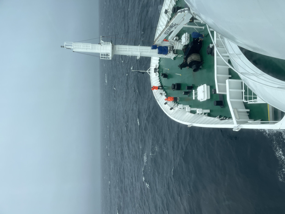 This high-angle view captures the bow section of a large ship navigating choppy, dark blue-grey waters under a heavily overcast sky. The image looks down from a high point on the vessel, likely from its superstructure or mast, towards the front of the ship.
The ship features a broad, green-painted main deck contrasted with white railings, fixtures, and part of its superstructure which curves prominently on the right side of the frame. At the very front, a tall, white mast structure stands, equipped with several platforms and possibly radar or communication devices. To the port side of the mast, a white crane with some blue coverings is visible; the text "B S O R M E C" is discernable on its side. Various other pieces of deck equipment, including hatches and bollards, are scattered across the deck.
Several individuals are visible on deck. Two people wearing bright orange life vests are positioned near the bow railings on the port side. Further aft and to starboard, one person in dark clothing is attending to equipment, while another person in blue stands near the crane.
The sea is rough, with visible waves and whitecaps, stretching into an indistinct, hazy light grey horizon. The sky merges seamlessly with the horizon due to thick cloud cover or mist, giving the scene a muted, cool color palette. No landmasses are visible, suggesting the ship is operating in open ocean. The daylight conditions are soft and diffused, characteristic of a heavily overcast day.