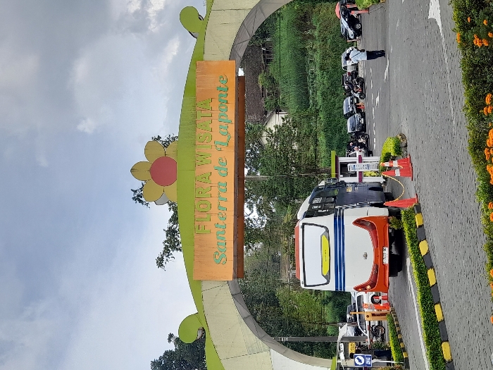 The image shows an entrance to the "FLORA WISATA Santerra de Laponte" attraction located in the city of Kecamatan Pujon, Indonesia, at coordinates (latitude: -7.854, longitude: 112.486). It's an archway made of yellow and orange painted brick with a large, yellow-and-green flower-shaped sign at the top and a sign below reading "FLORA WISATA Santerra de Laponte". A white bus with blue and orange stripes is parked inside the entrance, with the rear end facing the camera. The bus is in the foreground, with a few other vehicles parked behind it. A single man in a blue shirt is walking towards the right side of the image. The bus is parked on an asphalt lot, and there is a small row of orange traffic cones to the left and right of the bus. The surrounding area is lush, green vegetation. The sky is overcast, with a few clouds in the sky.