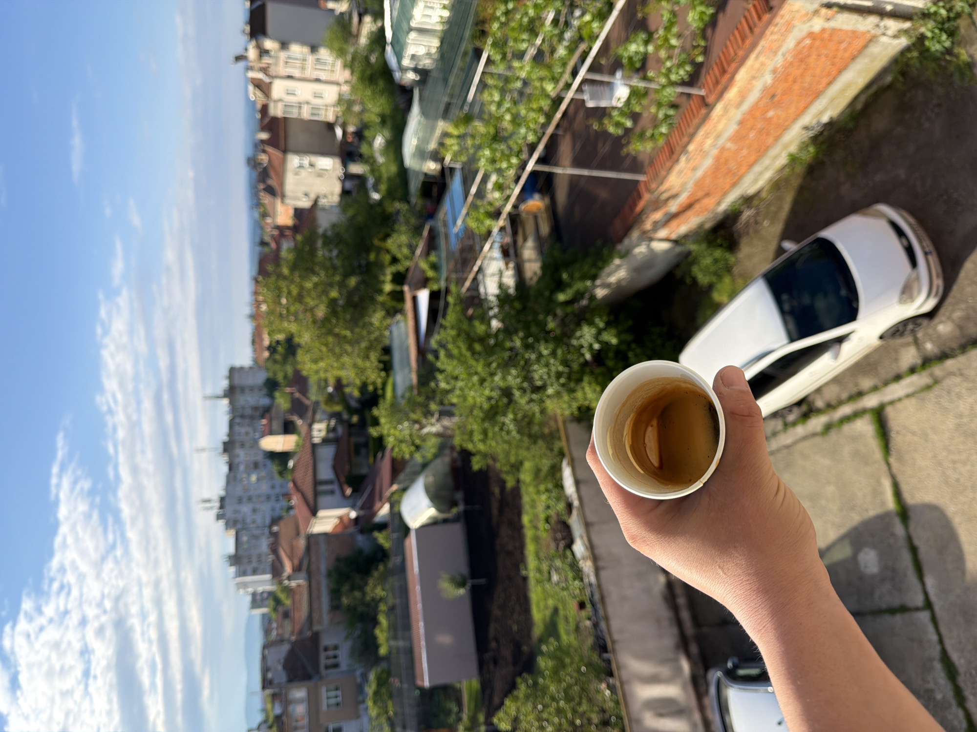 A person's hand holds a partially-filled white paper cup of coffee in the foreground. The background depicts an urban scene in Burgas, Bulgaria. A mix of residential buildings with varying heights and architectural styles are present. Green trees and vegetation fill the spaces between the buildings. The sky is blue with scattered white clouds. A white car is parked on a concrete surface near a brick wall. The image is taken from an elevated position, possibly a balcony or high window.