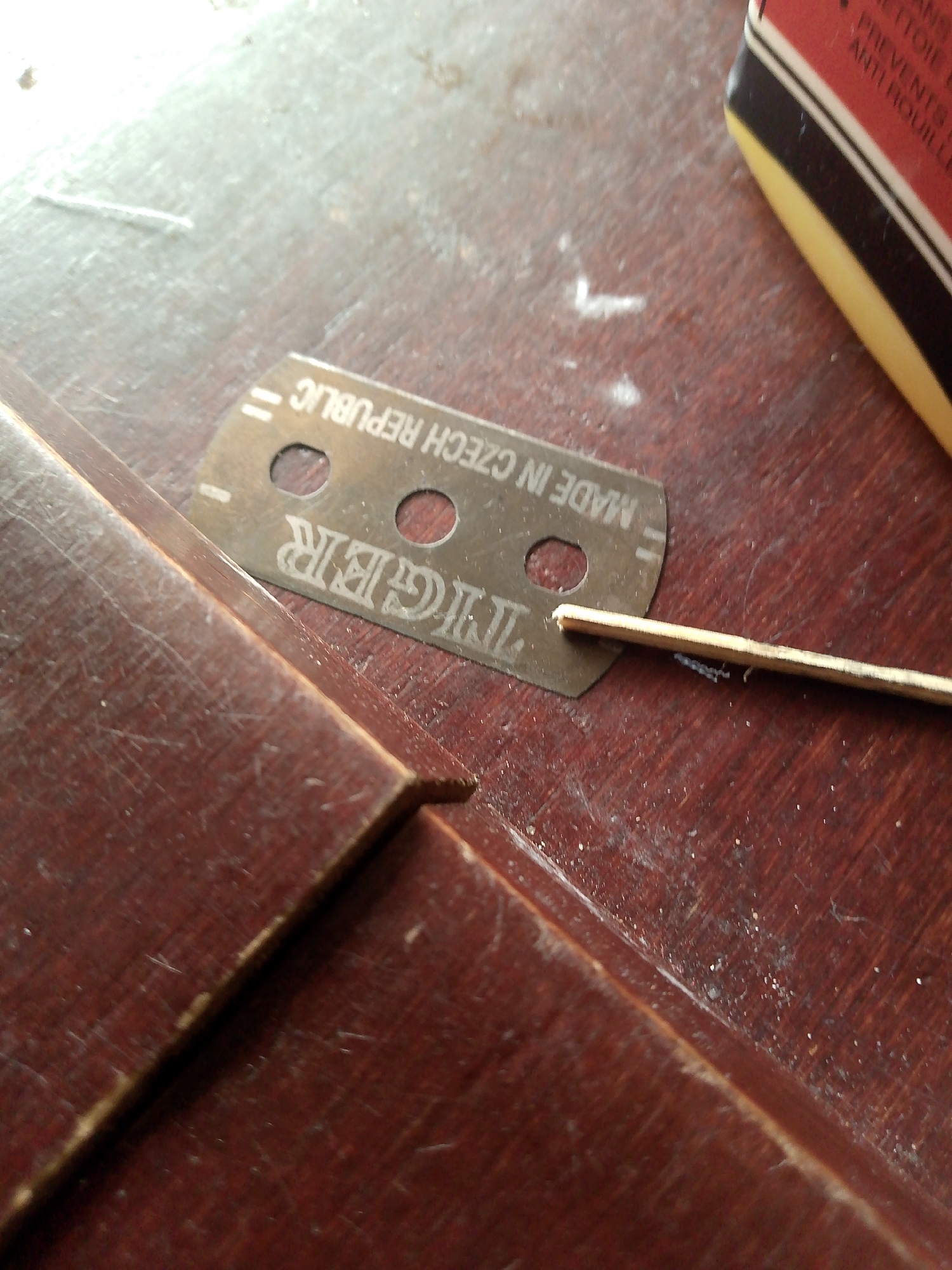 The image shows a close-up, overhead view of a wooden surface with a razor blade. The wooden surface appears to be a table or desk, with a dark brown, polished finish showing visible scratches and dust. A single-edge razor blade is placed horizontally on the surface, with the text "MADE IN CZECH REPUBLIC" and "TIGER" stamped on it. A thin, light-colored stick, possibly a wooden skewer or toothpick, is positioned beneath the razor blade, with its tip touching the blade's edge. In the upper right corner, a portion of a red and yellow box or packaging is visible. The scene is illuminated by overhead lighting, creating some highlights and shadows. There are no people present in the image. The image does not contain any information that would allow for its location to be determined in Jauro Yinu, Nigeria or any other specific city.