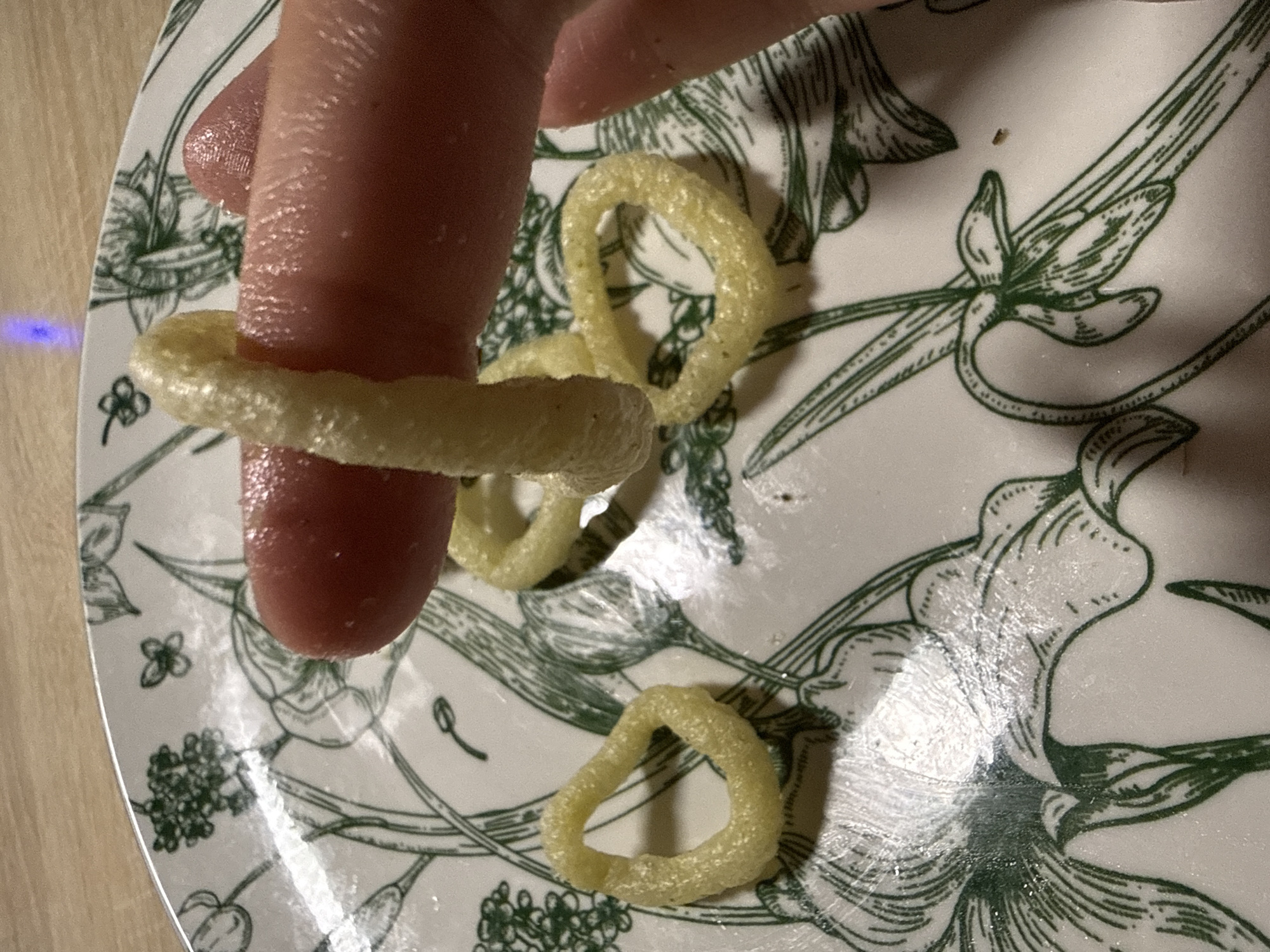 A person's index finger, coated in a glistening substance, is inserted into a yellow, ring-shaped snack food. The snack is textured with spiral ridges and appears somewhat brittle. The finger is positioned against a white plate decorated with green line drawings of botanical motifs, possibly irises and other flowers, and small butterfly-like insects. Several other similar snack rings are scattered across the plate. The background shows a wooden surface. The scene is illuminated by artificial light, which creates a slight blue reflection on the plate. There is no information to suggest this scene is located in Cheboksary, Russia.