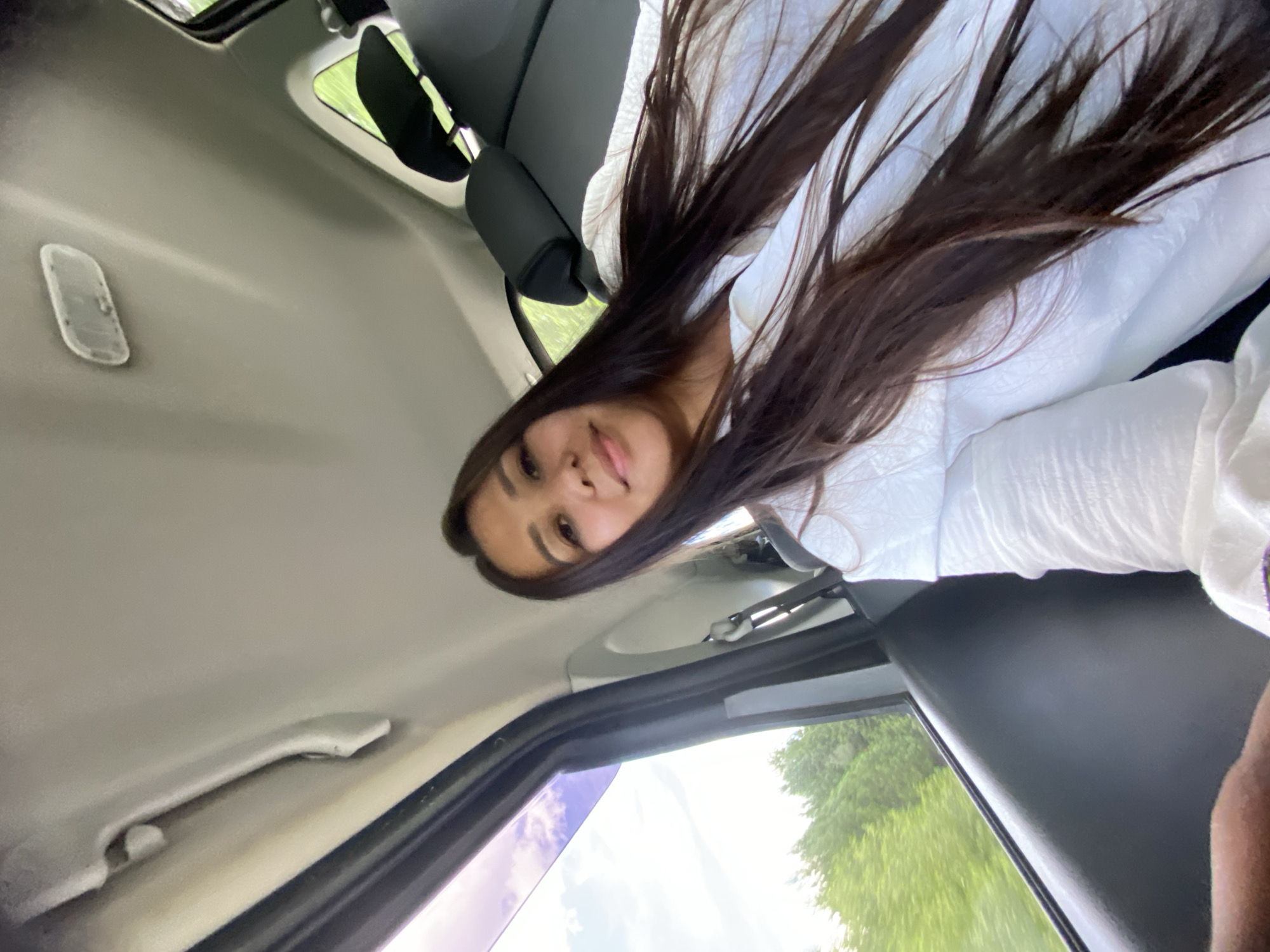 A young woman with long, dark hair is taking a selfie in the backseat of a car. She is wearing a white shirt. Through the open car window, a lush green forest is visible, suggesting a rural or suburban setting. The sky is partly cloudy. The car's interior, including the ceiling and headrest, is visible. The specific location in Zilair, Russia cannot be determined from the image alone.