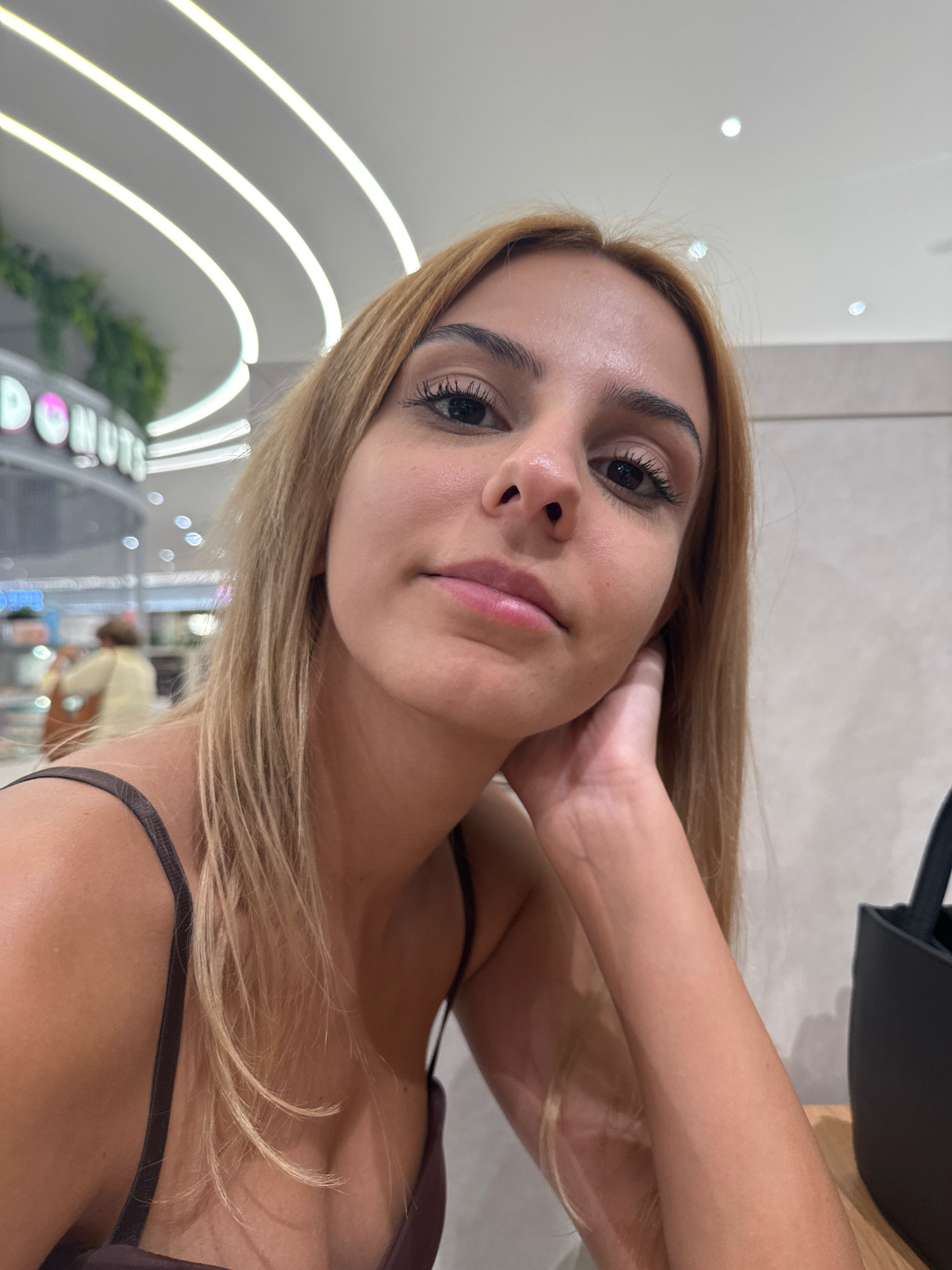In a dimly lit indoor setting, likely a mall or a commercial establishment with decorative lighting, a young woman of Caucasian ethnicity with blonde hair and light skin is captured in a close-up selfie. Her head is tilted to her right, with her left hand supporting her chin and cheek. She is wearing a dark-colored spaghetti strap top. The background features a sign that appears to read "DONUTS" in neon lettering, indicative of a food establishment. The ceiling has prominent curved white light fixtures. The overall atmosphere suggests a casual outing in an urban environment. No specific location in Sofia, Bulgaria, can be identified from the visual information provided.