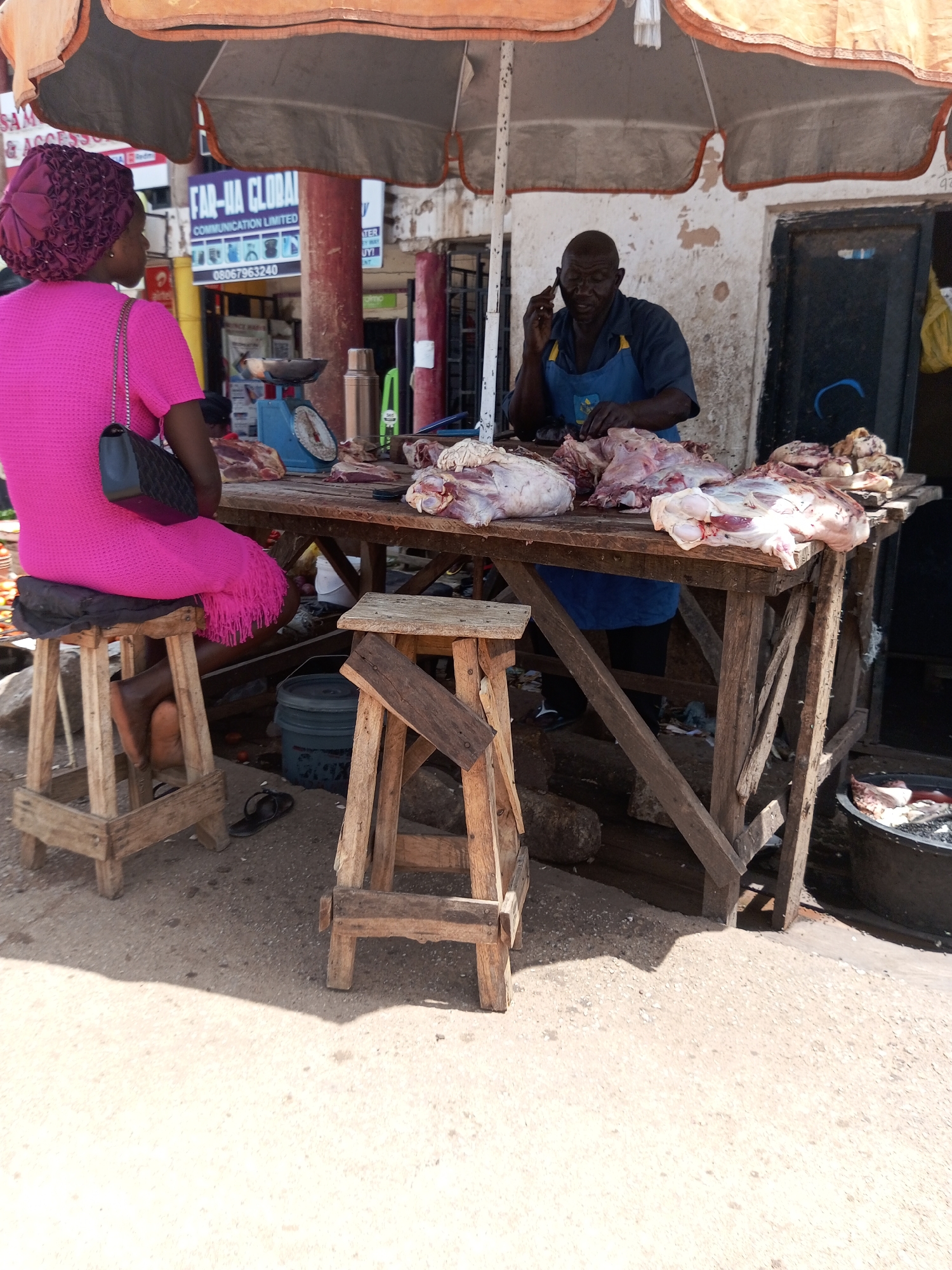 A butcher stands at a wooden stall in an outdoor market, talking on his phone while arranging raw meat. He is wearing a blue apron over a dark shirt. In front of him, a woman in a bright pink dress and headscarf is seated on a stool. The stall is covered by a large, open umbrella, providing shade. Various cuts of meat are displayed on the table, some on a blue scale. In the background, a building with peeling paint and a doorway are visible. The ground is sandy and dusty. There are other market stalls and structures in the distance, suggesting a busy marketplace. The lighting indicates it is daytime, likely mid-morning or afternoon. The scene appears to be in a warm climate. There is visible text on a sign in the background that reads "FAR-HA GLOBAL COMMUNICATION LIMITED" with a phone number and logos. Another sign appears to have text related to "SAM".