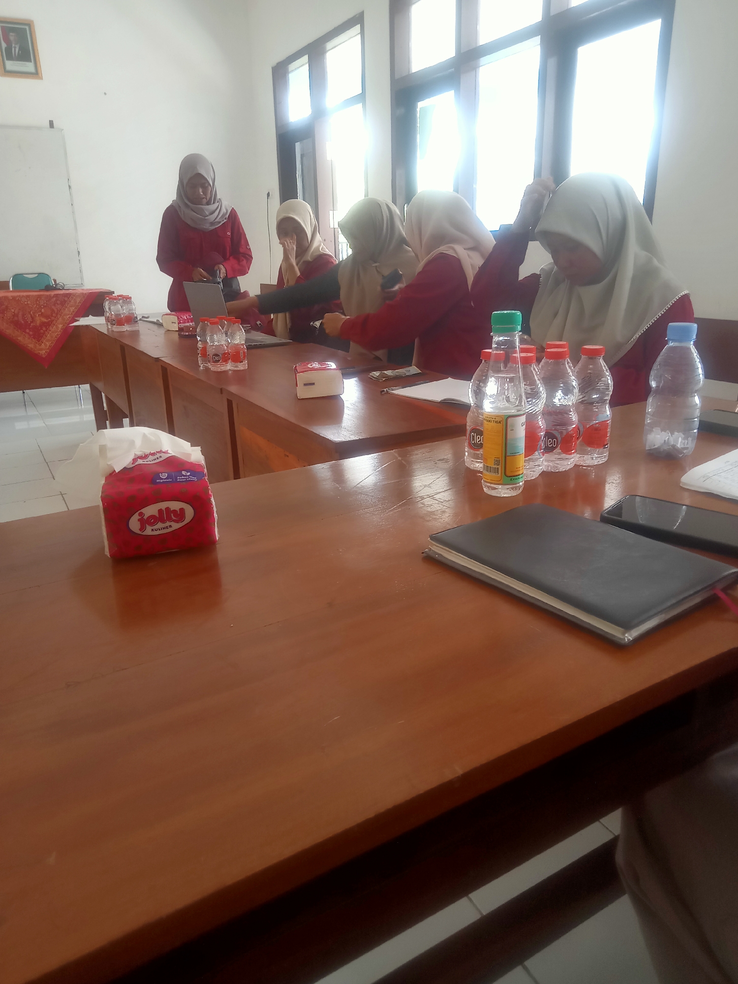 In a classroom setting in Pakel, Indonesia, at least four women wearing hijabs are seated at wooden desks, engaged in an activity that appears to be a meeting or presentation. One woman, standing at the front left, wears a red top and gray hijab and is looking at a laptop. To her right, another woman in a red top and cream hijab is also looking down. Further to the right, a woman in a red top and cream hijab is reaching up towards her head. The desks are arranged in rows, and the room features a whiteboard on the left wall and a framed portrait of a man in a suit above it. On the desks, several water bottles, a tissue box with a red Jolly Kuliner design, a notebook, and a smartphone are visible. Sunlight streams in through large windows on the right side of the room, illuminating the scene. The atmosphere suggests an educational or administrative gathering.