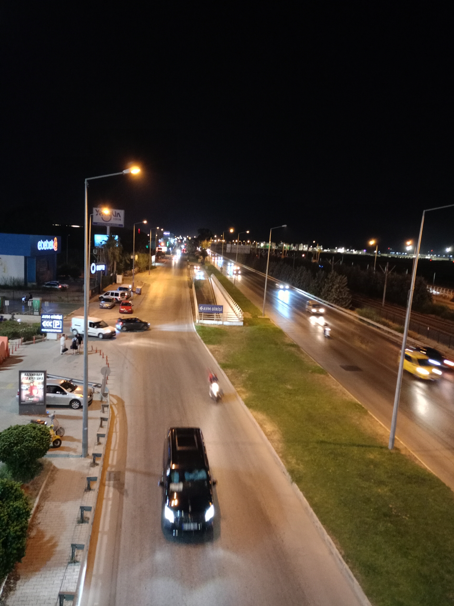 The image captures a nighttime street scene in Kepez, Turkey, with a focus on a multi-lane road and surrounding urban elements. A black van is prominently positioned in the foreground, driving on the right lane of the road. Its headlights are illuminated, indicating movement. To the right of the van, a yellow vehicle, possibly a taxi, is visible, also in motion. The road is wet, reflecting the streetlights and vehicle lights, suggesting recent rain or damp conditions.
On the left side of the road, a blue building with a white sign that reads "ebbek" is partially visible, likely a commercial establishment. Adjacent to it, a white van and several cars are parked. There are also pedestrians near the parked vehicles, some carrying bags, suggesting activity around the commercial area. Streetlights are illuminating the scene, casting a warm glow.
Further down the road, more vehicles are seen, with their lights creating streaks due to the long exposure or motion blur. A grassy median separates the lanes of the main road, and further to the right, there's another lane of traffic and more grassy areas. In the distance, city lights and structures are visible against the dark night sky. A signpost with the text "AVM GİRİŞİ" and arrows indicates an entrance to a shopping mall. The overall impression is a busy urban environment at night, with ongoing traffic and commercial activity.
