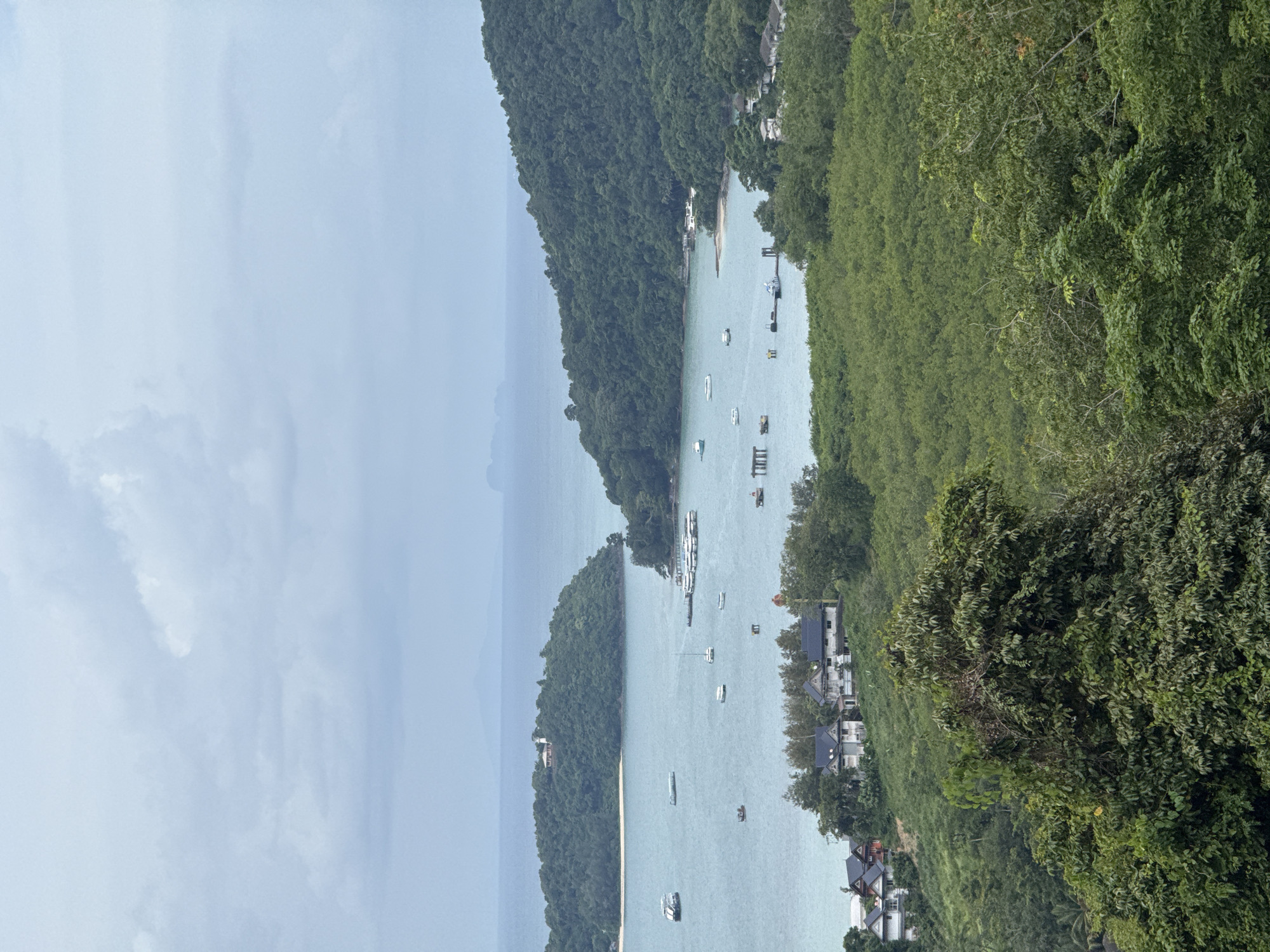 The image depicts a scenic overview of a bay with numerous boats docked at a pier. Lush green hills covered in dense vegetation slope down to the water. Several buildings, possibly residential homes, are nestled on the hillsides overlooking the bay. The water is a calm, blue-green hue, and the sky is overcast with soft, grey clouds. In the distance, more islands are visible across the horizon. The scene is tranquil and natural, suggesting a tropical coastal location. This view is likely from a vantage point within or near the city of Wichit, Thailand, known for its beautiful coastlines and islands.