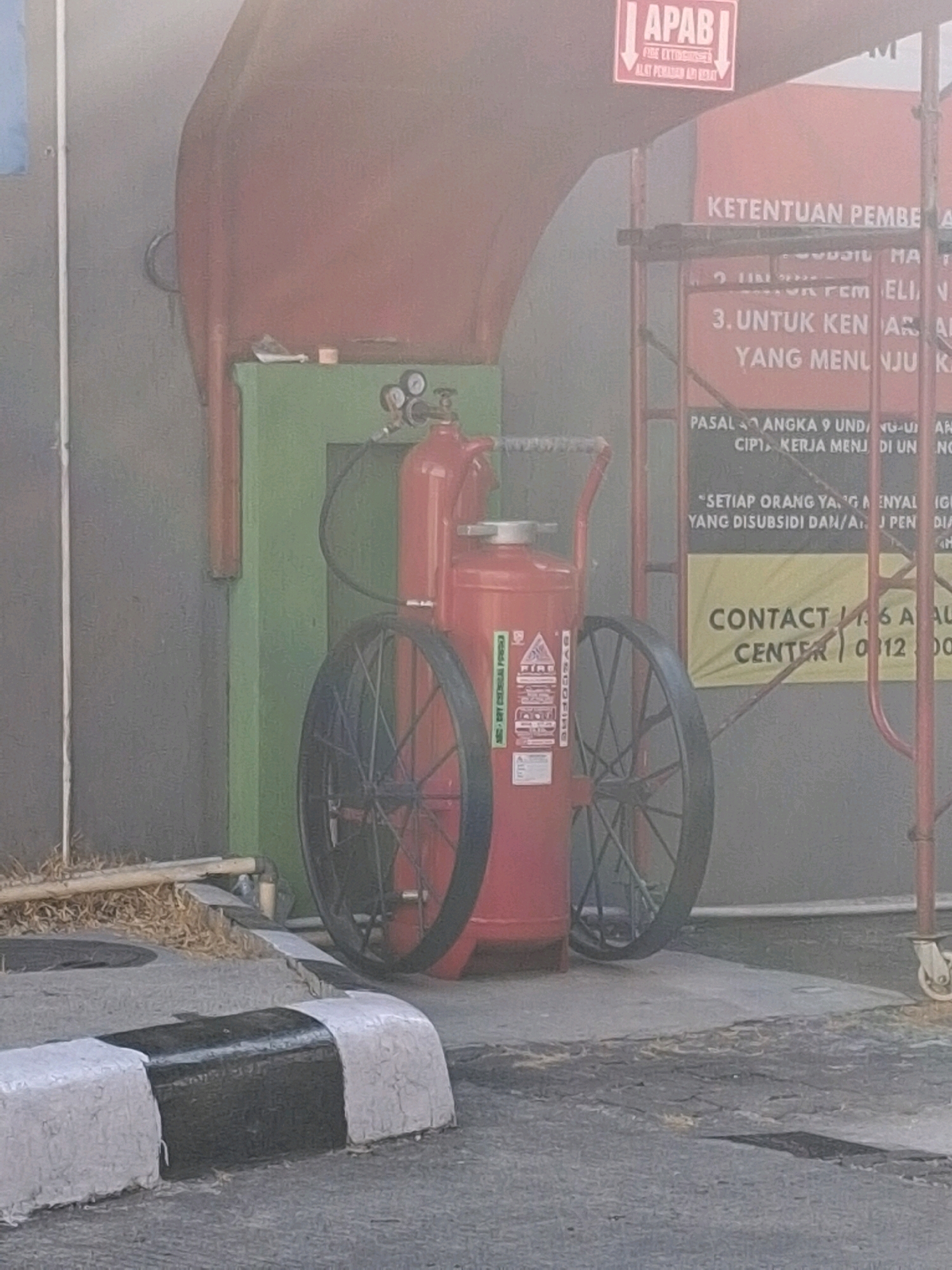 A red fire extinguisher on wheels is positioned outdoors in Surakarta, Indonesia, near a building with green and red elements. The fire extinguisher has large spoked wheels and a handle. There are signs visible on the building, one with "APAB" and another that appears to be a list of regulations. In the foreground, a curb painted with black and white stripes is visible, and the ground is paved. Some dry grass or straw is scattered on the ground. The overall scene suggests an industrial or public area, possibly a facility that requires fire safety equipment.