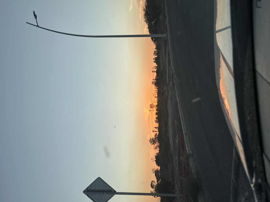 An outdoor scene shows a roadside view at sunset, likely captured from inside a vehicle. The sky blends from blue to orange near the horizon, with trees silhouetted in the background. A tall street light pole and a diamond-shaped road sign stand beside the road. Part of a car's interior, including the window frame and a hazy reflection of the sunset, is visible on the right side of the frame.