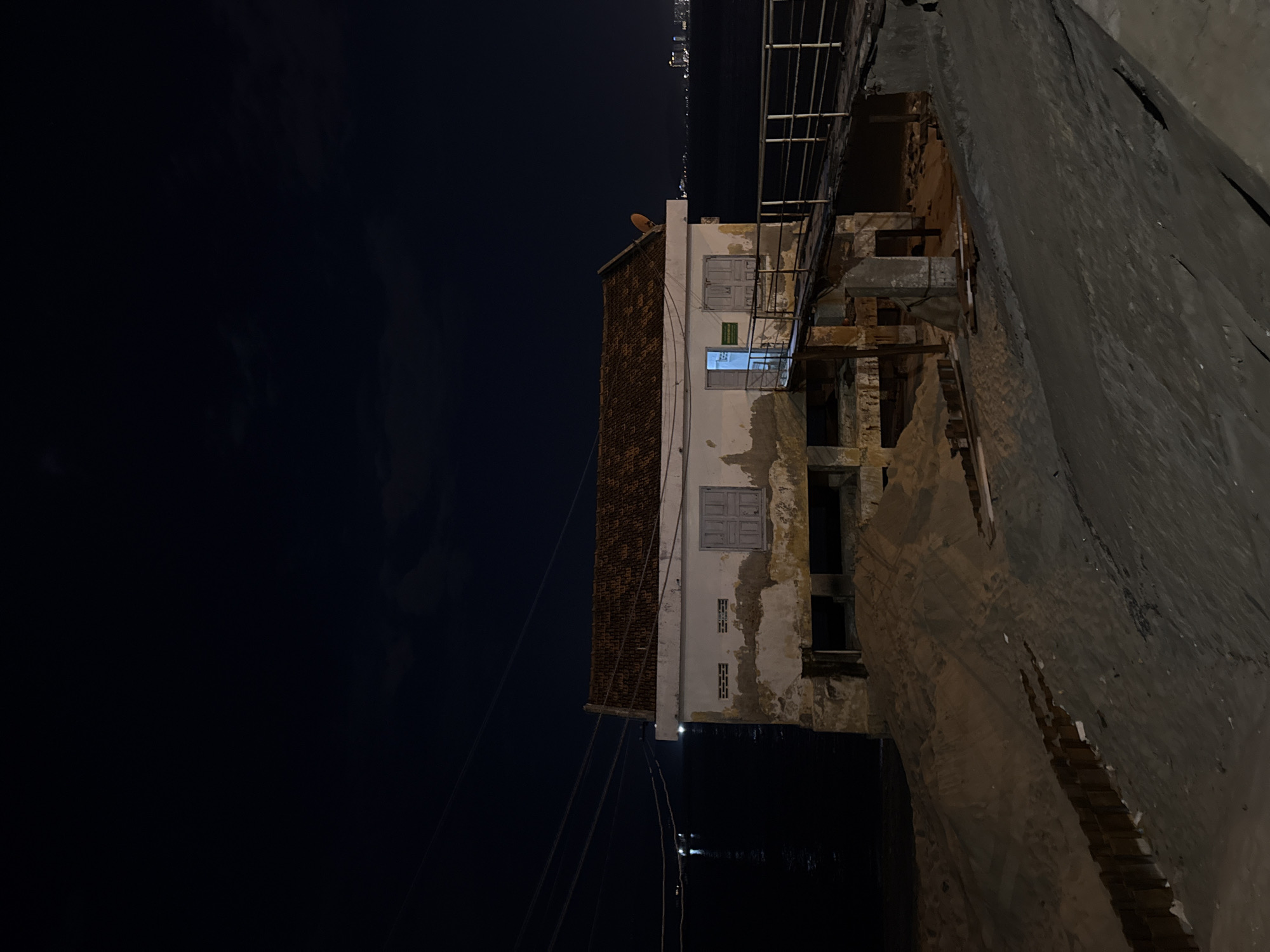 The image captures a nighttime view of an old, dilapidated house situated near the coast in Nha Trang, Vietnam. The house, with a tiled roof and weathered walls, appears to be elevated on a concrete foundation. A wooden walkway with railings leads to the entrance, where the door is ajar and a faint light emanates from within. The surrounding area is characterized by sand and debris, suggesting a construction or renovation site, or perhaps the effects of coastal erosion. In the background, the dark expanse of the ocean stretches out, with distant city lights visible on the horizon. Several power lines crisscross the sky above the water. The overall atmosphere is one of decay and abandonment, illuminated by the artificial light of the night.