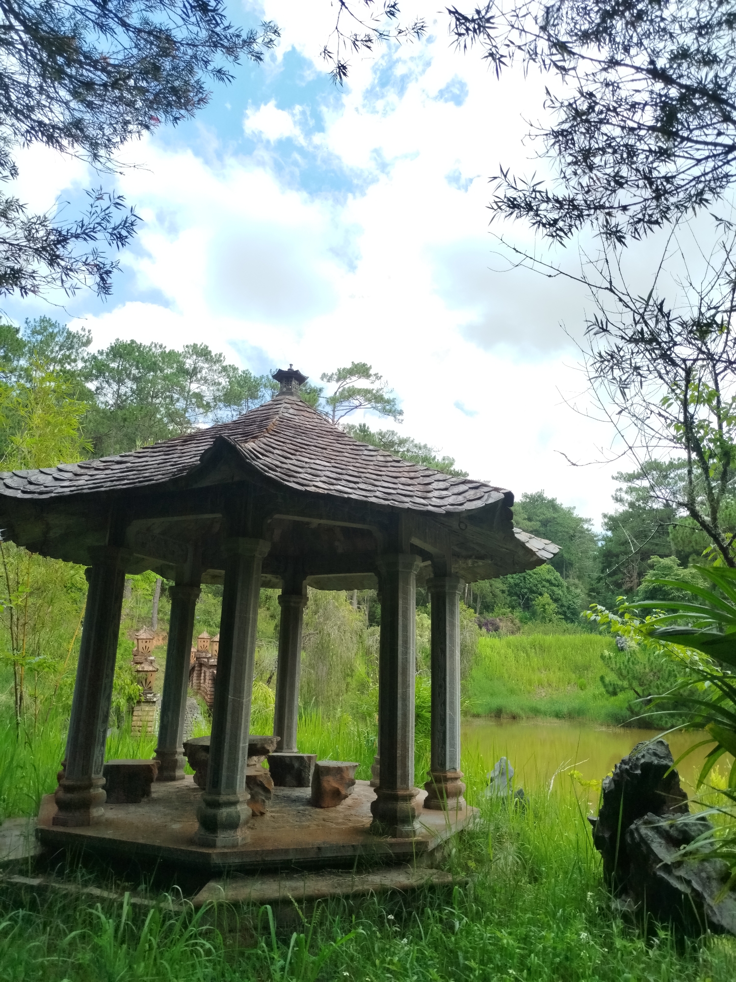 The image captures a pavilion in Cam Ly - Da Lat, Vietnam. The pavilion features a dark tiled roof and stone columns. Inside, there are stone blocks that appear to serve as seats. It is situated near a body of water, likely a pond or a lake, with greenery surrounding it. The sky is visible above, partially obscured by tree branches.