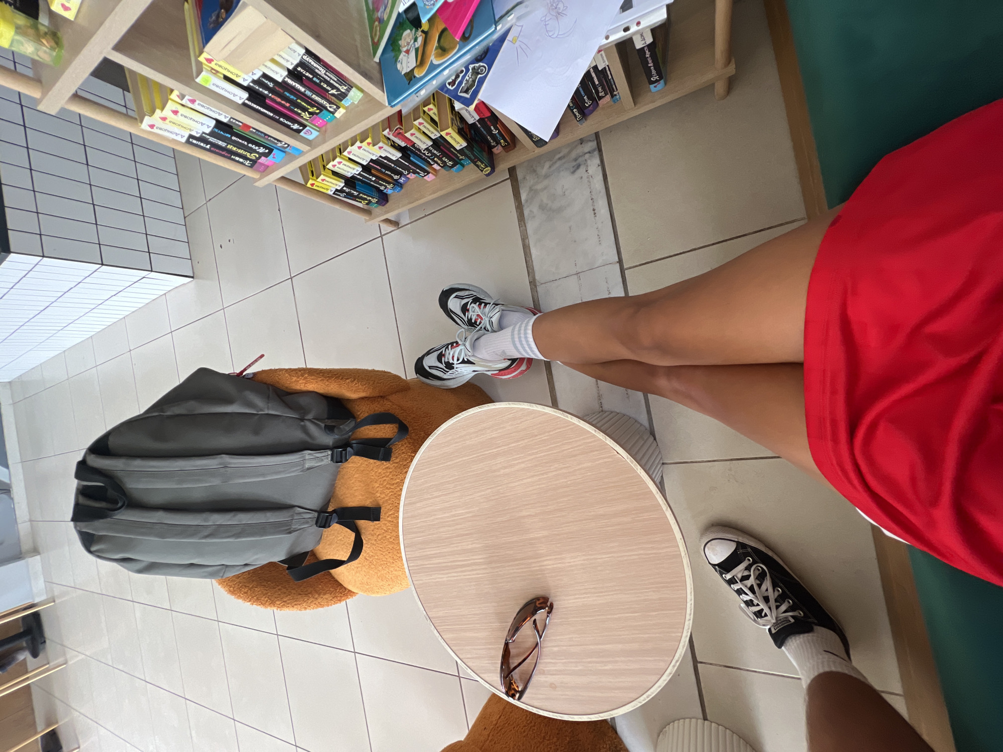 A first-person perspective shot shows a person's legs, wearing white socks and athletic shoes, crossed over their legs. The person is sitting in an orange chair with a grey backpack on it. Next to the chair is a light wood table with a pair of sunglasses on it. A bookshelf filled with books is visible to the right of the image. The scene appears to be indoors, possibly a cafe or library, in Antalya, Turkey.