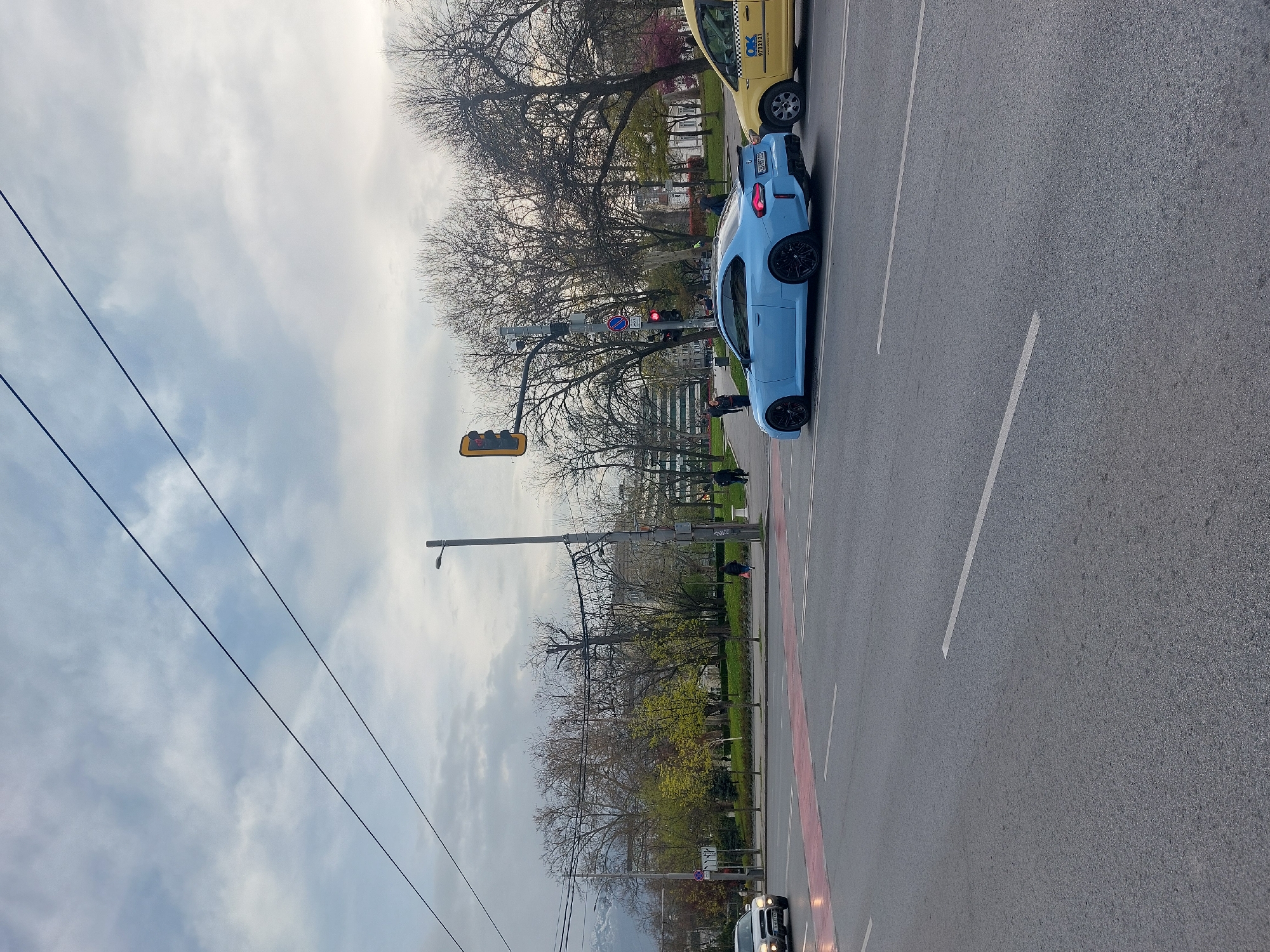 This image depicts a daytime urban street scene under an overcast sky. The primary subject is a light blue sports car, possibly a BMW M2, stopped or parked on the right side of a multi-lane asphalt road. Its rear design, including a spoiler and black wheels, is clearly visible, along with a European license plate reading "BG B0001AM". A yellow taxi cab is partially visible next to the blue car, displaying "OK" and "97321" on its side. Another white car is partially visible in the bottom right corner.
On the left side of the road, a park area features numerous trees, some with bare branches and others beginning to sprout green leaves, suggesting late winter or early spring. Several pedestrians are scattered on the sidewalks and within the park. Traffic lights are visible, with one showing red and another displaying yellow. A street sign, a red circle with a white horizontal bar, indicates "No Entry." Utility poles with overhead wires crisscross the upper portion of the frame. The road itself features white lane markings and a distinct red-painted section, possibly a bus lane or bike lane. The scene captures a typical, unextraordinary moment in a European city.