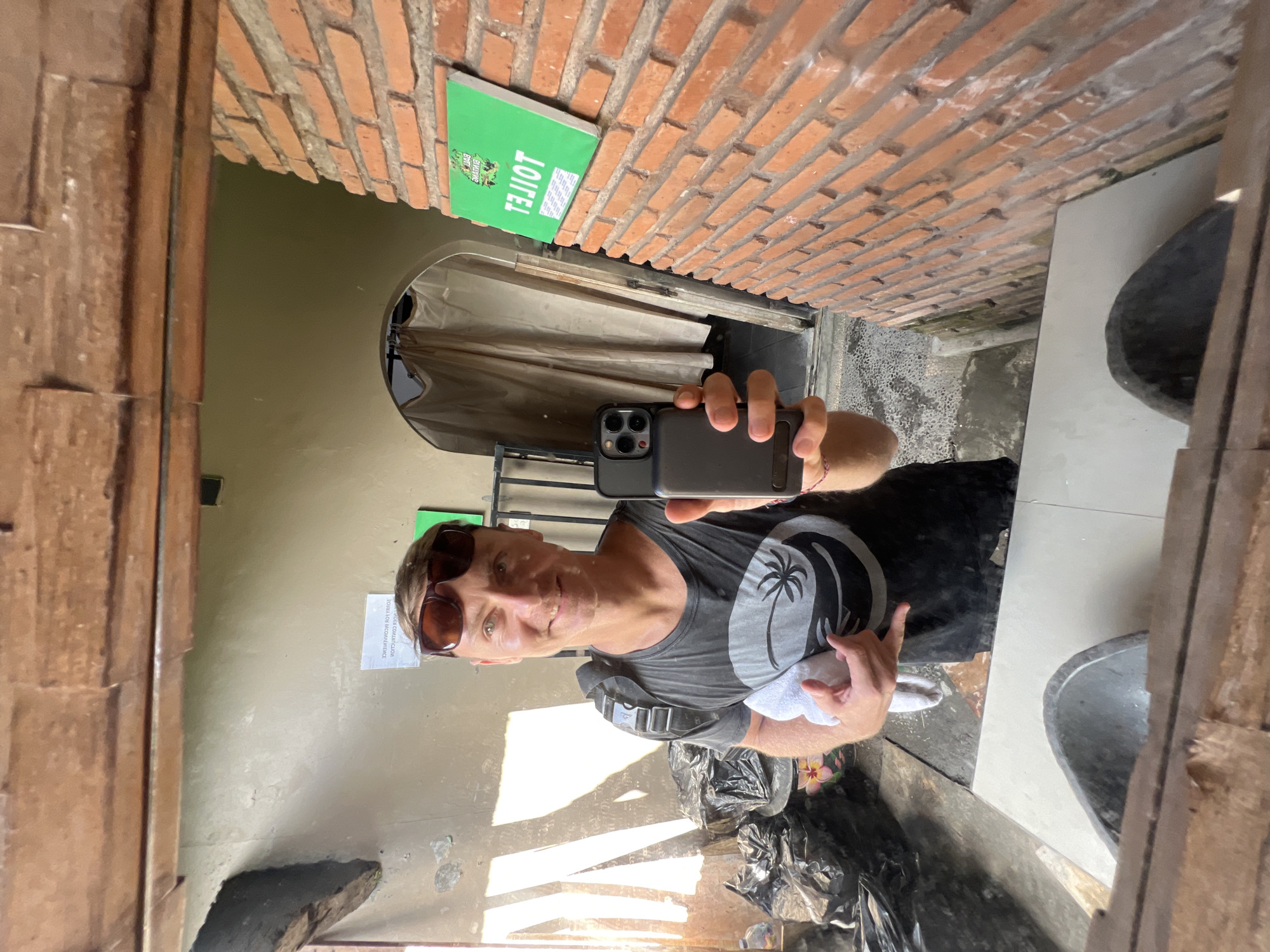 A man is reflected in a mirror, holding a phone to take a selfie in a room with a toilet sign on a brick wall and sink. He is wearing sunglasses and a t-shirt. There is a trash can nearby. The location appears to be in the Ubud District, Indonesia.