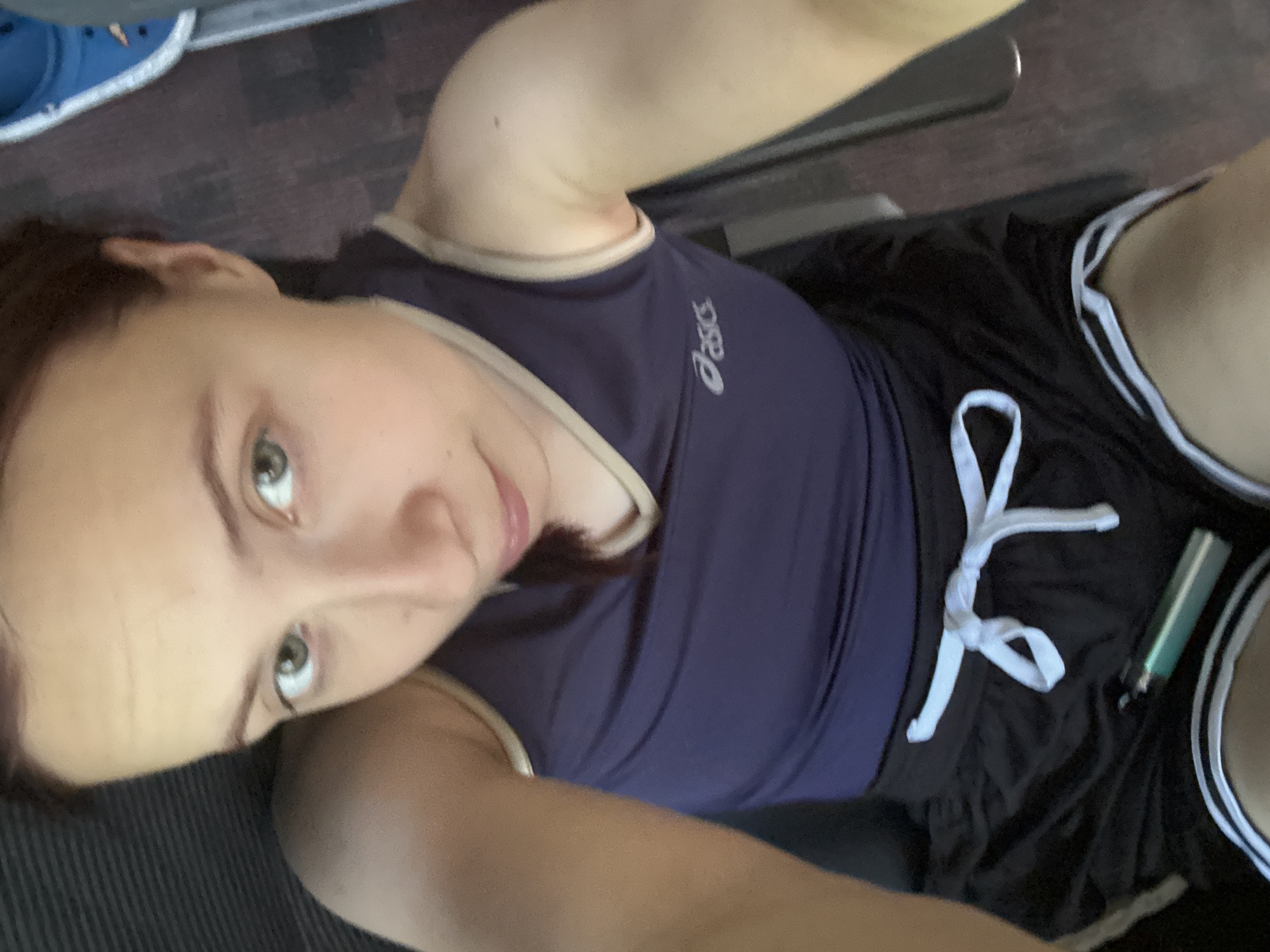 A selfie shows a light-skinned person with brown hair and green eyes wearing a navy blue Asics tank top and black shorts with a white drawstring. A green vape pen is visible on the right side of the shorts. The background appears to be a treadmill with blue shoes visible in the upper left corner. The location is unknown, but the user requested that it be placed in the city of Старое Мартьяново, Russia.