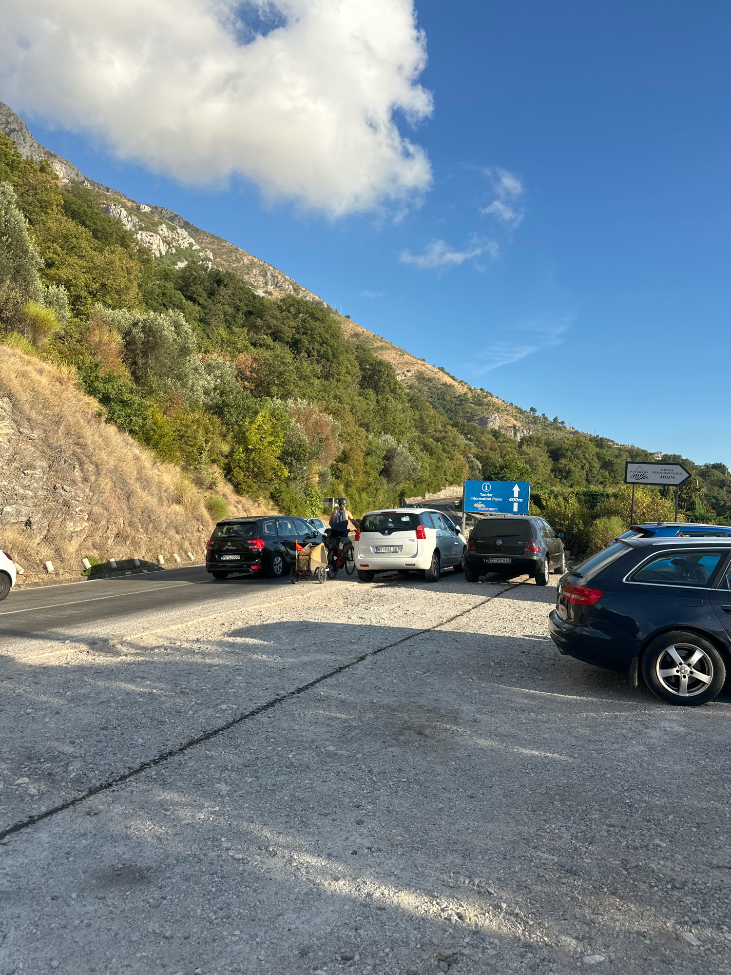 The image shows a parking area next to a winding road, with a steep, vegetated mountainside rising behind it. Several cars are parked, including a black hatchback, a white minivan, a dark grey hatchback, and a dark blue station wagon. A person is standing next to the black hatchback, next to a bicycle with a small trailer attached. The scene appears to be set on a sunny day with some clouds in a clear blue sky.
Two road signs are visible. One is a blue sign with white text and an icon of a person with a circle and arrow, indicating "Tourist Information Point 400m". The other is a white sign with black text and an arrow pointing right, which reads "LOUNGE PANORAMA RESTAURANT BAR 100m".
The overall impression is of a scenic stopover point, likely a rest area or a place to access local attractions, given the presence of the tourist information and restaurant signs. The time of day appears to be late morning or early afternoon, judging by the shadows. The environment suggests a Mediterranean or Balkan coastal region, consistent with the provided location context of Drobnići, Montenegro, known for its coastal roads and mountainous terrain. The activity appears to be a pause in travel, with individuals stopping to rest or consult directions.