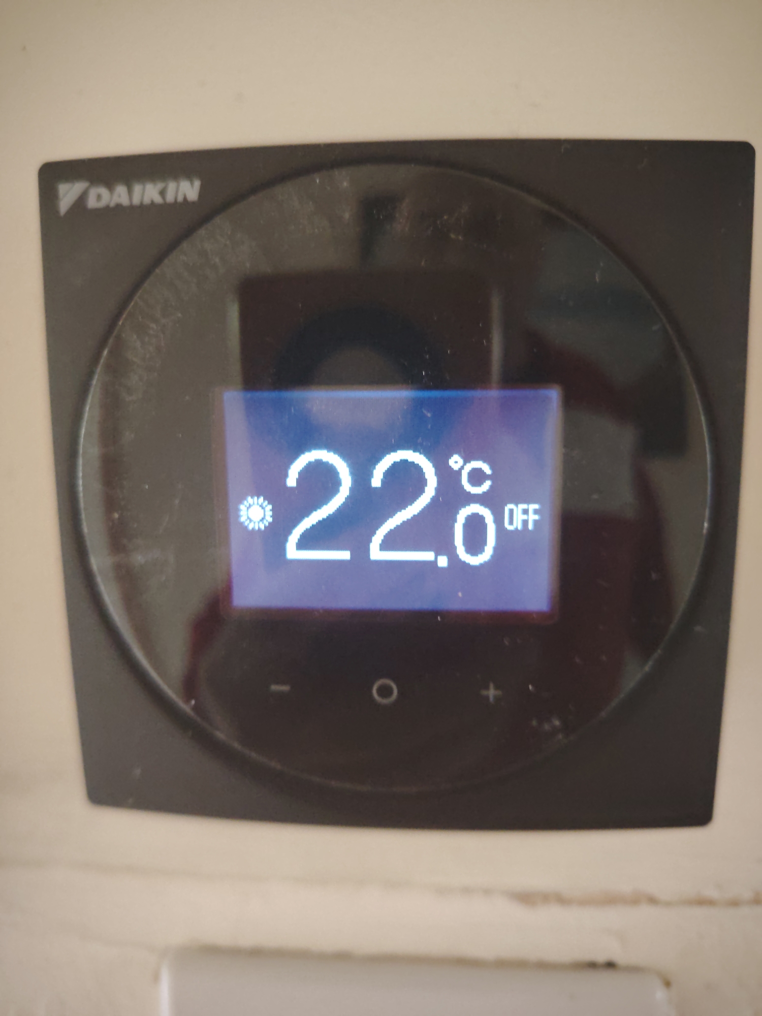 A close-up, slightly angled shot shows a Daikin thermostat or control panel mounted on a light-colored, possibly off-white or beige wall. The device is predominantly black with a circular, glossy face and a rectangular digital display at its center.
The brand name "DAIKIN" is visible in white text at the top left of the device's circular face. The screen is illuminated with blue backlighting, showing white text and numbers. A sun-like icon, typically indicating a heating mode or desired heating temperature, is displayed on the left side of the screen. To its right, the temperature "22.0" is clearly visible, followed by the degree Celsius symbol "°C". Further to the right, the word "OFF" is displayed, indicating that the heating or cooling system controlled by the thermostat is currently inactive.
Below the screen, along the bottom edge of the circular face, three touch controls or buttons are visible: a minus sign ("-"), a circle ("O", likely for power or mode selection), and a plus sign ("+"). The glossy surface of the device and screen shows reflections, suggesting ambient light in the room, and there are also visible smudges and dust particles on the surface. No people are present in the image. The exact time of day or weather conditions are not discernible from the photograph.