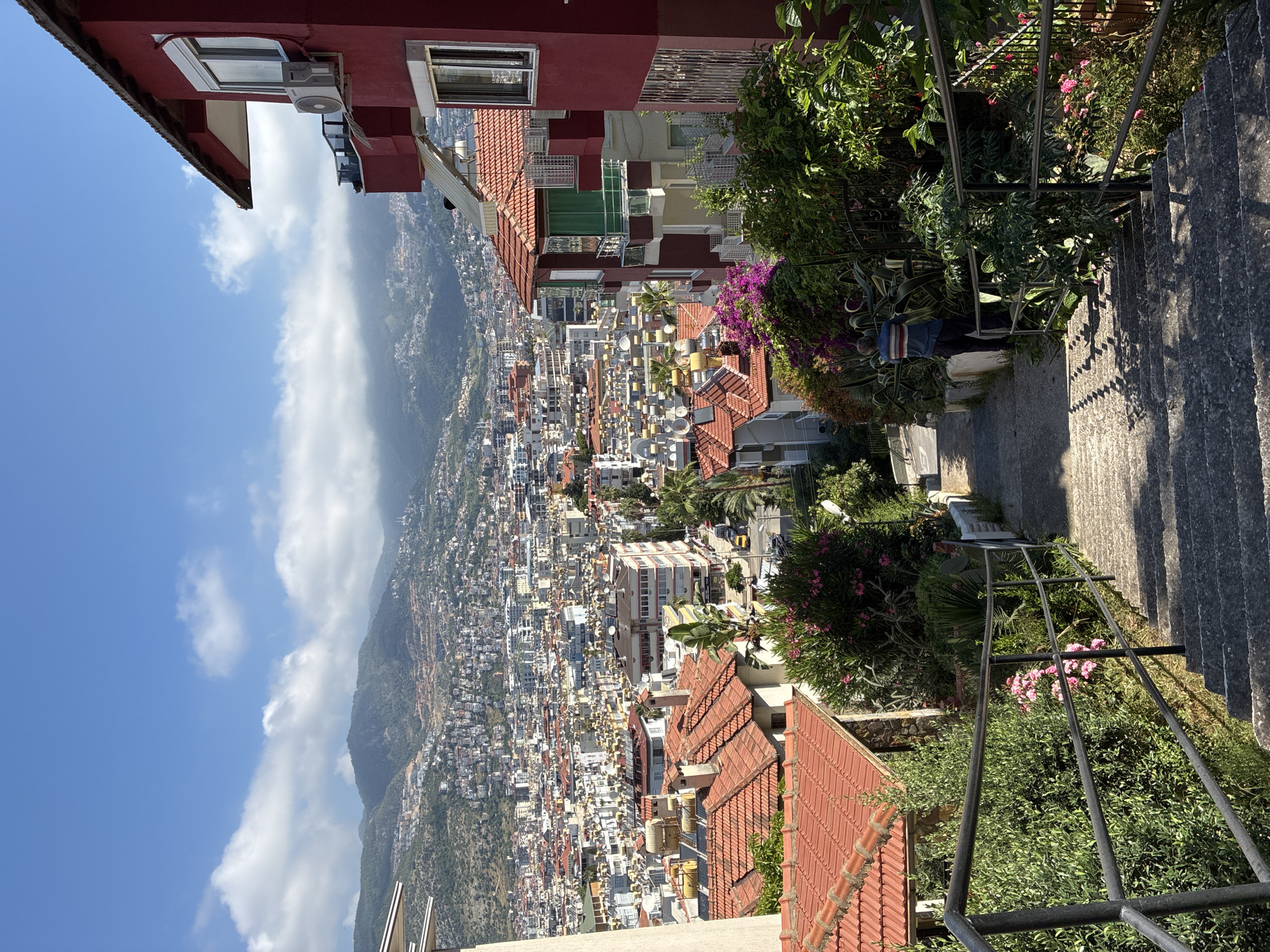 This image presents a panoramic, high-angle view of a sprawling city, likely taken from a steep hillside overlooking the urban expanse. The city is nestled against a backdrop of imposing green mountains, stretching under a clear blue sky dotted with scattered white clouds.
In the immediate foreground, to the left, is a vibrant red building with a terracotta-tiled roof, featuring a window and an external air conditioning unit. To the right, a concrete staircase with a dark metal handrail descends downwards, bordered by lush gardens. These gardens burst with greenery, including large succulents, leafy shrubs, and flowering plants with pink and purple blossoms, possibly bougainvillea. An indistinct figure, wearing a blue and white striped top, is vaguely visible near the stairs.
The middle and background reveal a dense urban landscape characterized by countless buildings, many featuring distinctive red-tiled roofs and light-colored facades. Numerous satellite dishes are visible on the rooftops, indicating residential areas. Interspersed among the buildings are various trees, including palm trees, adding pockets of natural greenery. The cityscape appears to climb the lower slopes of the distant mountains, which are covered in vegetation.
The bright sunlight suggests it is daytime, casting clear shadows, particularly on the staircase. The overall scene conveys the impression of an extensive and vibrant city integrated into a dramatic mountainous coastal environment, aligning with the provided context of Alanya, Turkey. There is no visible text in the image.