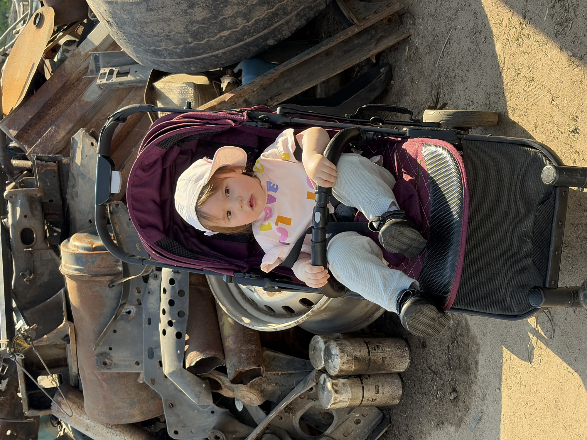 A baby wearing a white sun hat, a pink shirt with colorful letters, and light blue pants is sitting in a purple and black stroller. The stroller is surrounded by a pile of scrap metal, including tires, wheels, pipes, and various rusty metal parts. The ground appears to be dirt or gravel. The scene is lit by sunlight. The image was taken in Chaikovsky, Russia.