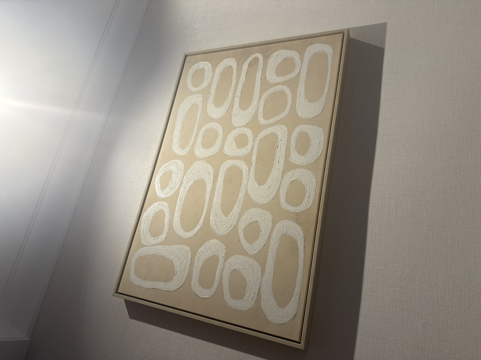 A framed abstract painting hangs on a textured, neutral-colored wall. The painting features a pattern of irregular, oblong shapes in a textured white paint against a light tan background. The shapes are outlined with a thick, impasto stroke, creating a sense of depth and tactility. The frame is thin and light-colored, appearing to be wood.
To the left of the painting, a bright light source casts a dramatic shadow diagonally across the wall and the painting itself. This light appears to be coming from an unseen window or lamp, and its intensity suggests it is daytime, possibly morning or late afternoon given the angle of the light and shadow. The wall texture is visible and appears to be a woven fabric or linen wallpaper, common in modern interior design. The overall scene is minimalist and contemporary, evoking a sense of calm and artistic contemplation. The location context of South Jakarta, Indonesia, doesn't provide specific visual cues in this image. No text is visible in the image.