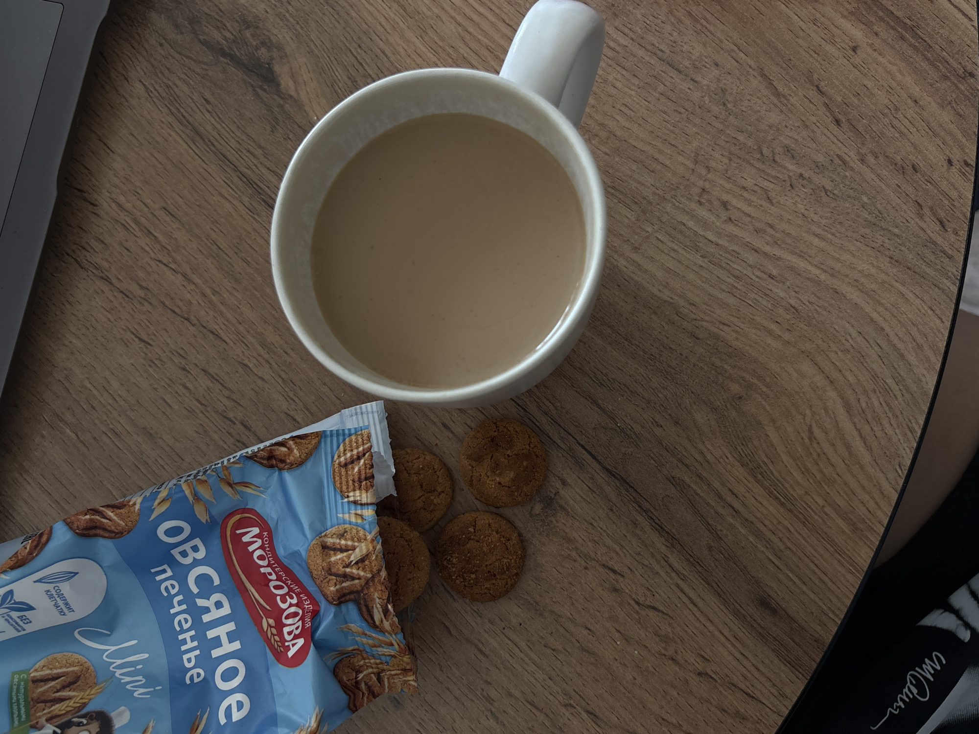 The image shows a bag of oat cookies and a mug of coffee on a wooden table. The bag is partially open, and several cookies have fallen out. The mug is white and filled with coffee. The table is made of wood with a visible grain pattern. In the bottom left corner of the image, there is a black shoe with a white logo. The image is taken from a top-down perspective. The scene is indoors, likely in a home or office. There is no information in the image that indicates the location of Rostov-on-Don, Russia.