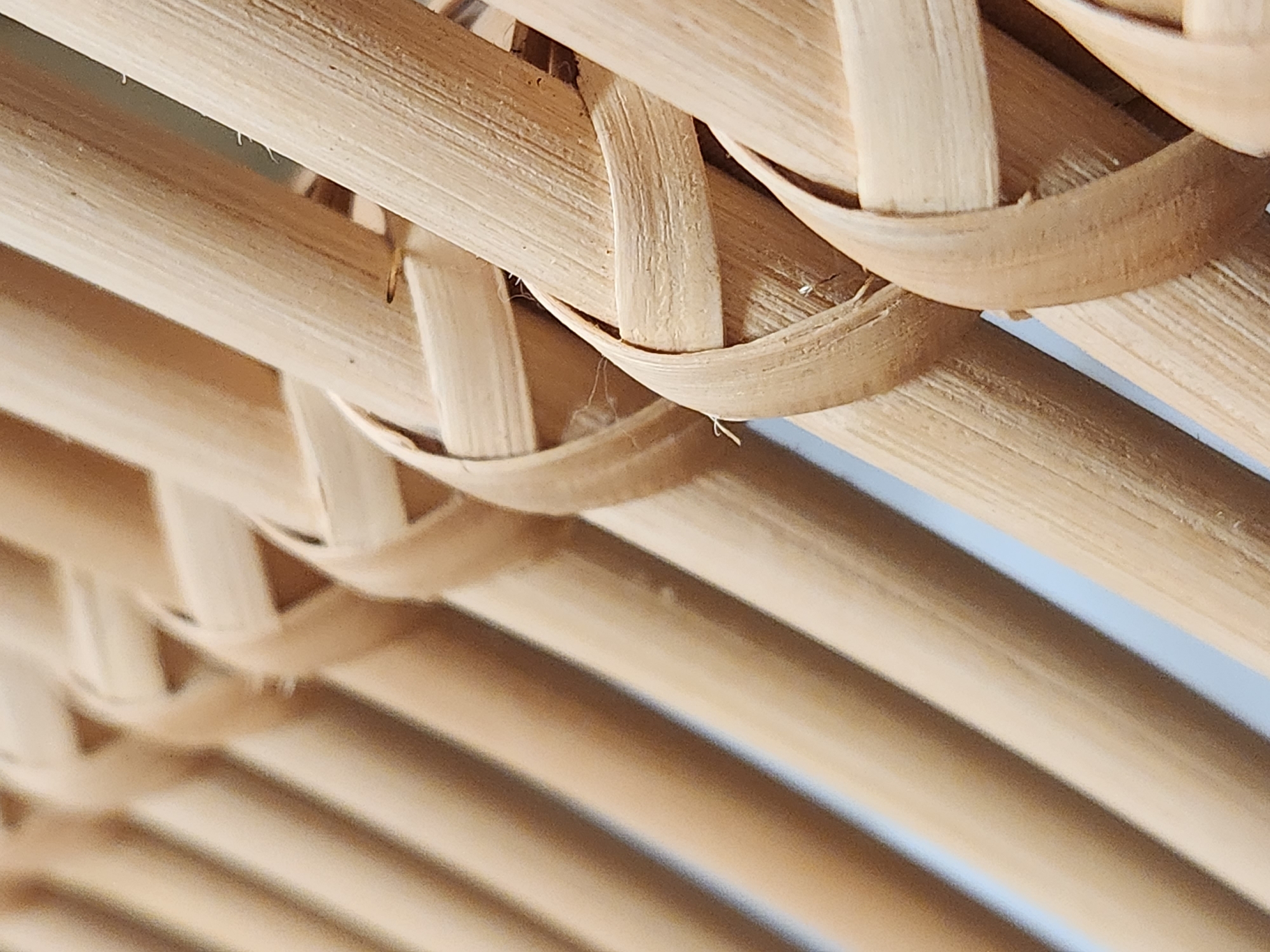 The image shows a close-up of a woven material, likely bamboo or rattan, forming a textured surface. The natural fibers are light brown and are interlaced in a repeating pattern, creating a basket-like construction. Some strands are thicker and form the base, while thinner, more flexible strands are woven over and under them to create the decorative and structural weave. The lighting suggests an indoor or shaded outdoor setting, with soft illumination highlighting the texture and slight variations in the color of the material. The background is out of focus, appearing as a soft blur of light blue and brown, suggesting it might be another surface or the sky. There are no people, text, or specific location cues visible within the frame. The image focuses entirely on the material and its woven structure, emphasizing the craftsmanship and natural beauty of the material.
