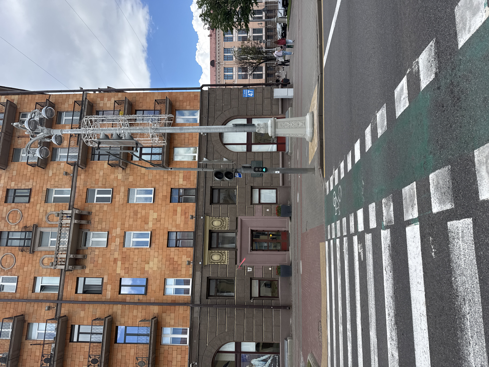 The image shows a view of the intersection of Nezavisimosti Avenue and Komsomolskaya Street in Minsk, Belarus. On the left is a multi-story brick building with numerous windows and balconies. In the foreground, a pedestrian crosswalk is visible, marked with white stripes and green paint indicating a bike lane. A traffic light and a street lamp are positioned near the intersection. Across Nezavisimosti Avenue, another building with a pink facade and numerous windows is visible, with trees and parked cars in front of it. Several people are walking on the sidewalk. The sky is blue with scattered clouds.