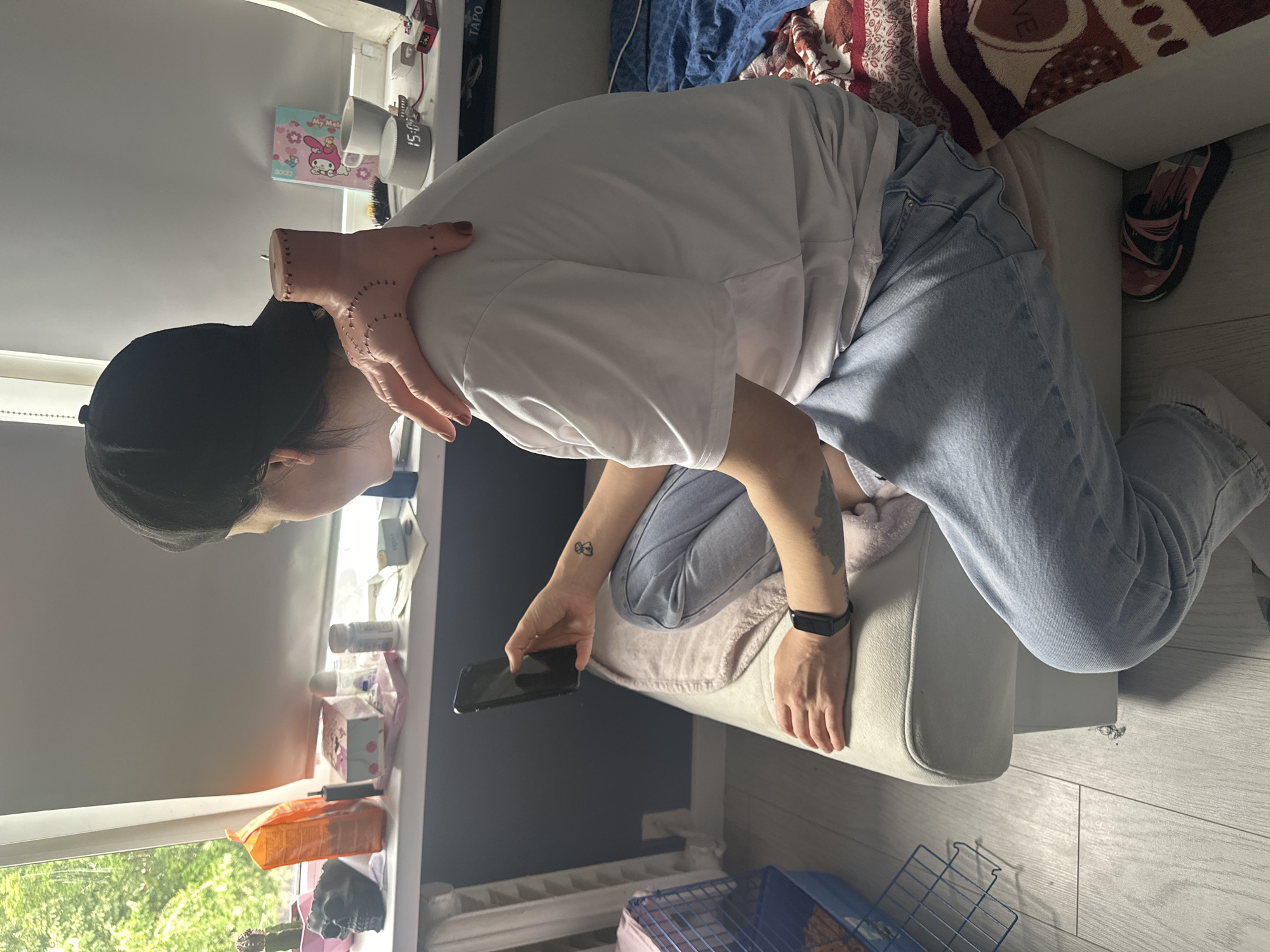 A person wearing a black cap, white t-shirt, and light blue jeans is kneeling on a white upholstered ottoman, holding a black smartphone in their left hand. The person has a tattoo on their left wrist and another on their left forearm. The background shows a window sill with various objects on it, including containers, a clock, and a My Melody box. A blue and pink cage is visible on the floor. A bed with a patterned blanket is partially seen in the upper right corner. The flooring is light gray wood grain. The location is in a residential setting, possibly in the city of Stavropol, Russia.