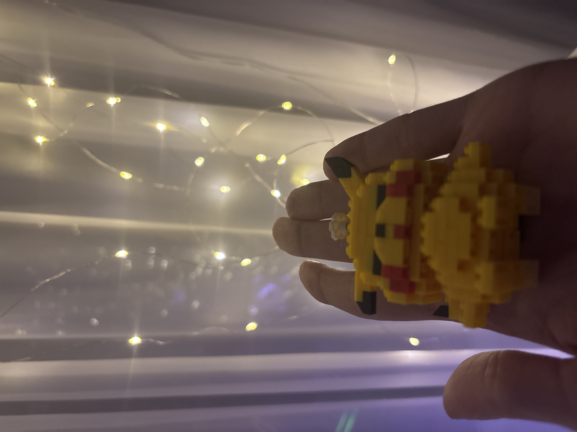 A close-up, medium shot shows a hand holding a pixelated yellow Pikachu figurine. The hand is open, palm up, and the figurine is resting on it. The Pikachu is facing forward and is primarily yellow with red cheeks and black accents. In the background, a sheer white curtain is visible, draped with thin strands of small, warm-toned LED lights. The lights are out of focus, creating a bokeh effect. The lighting is dim, with the warm glow of the fairy lights illuminating the scene. The overall impression is cozy and whimsical, with the figurine and lights creating a magical atmosphere. The location of the scene is Unknown, Unknown.