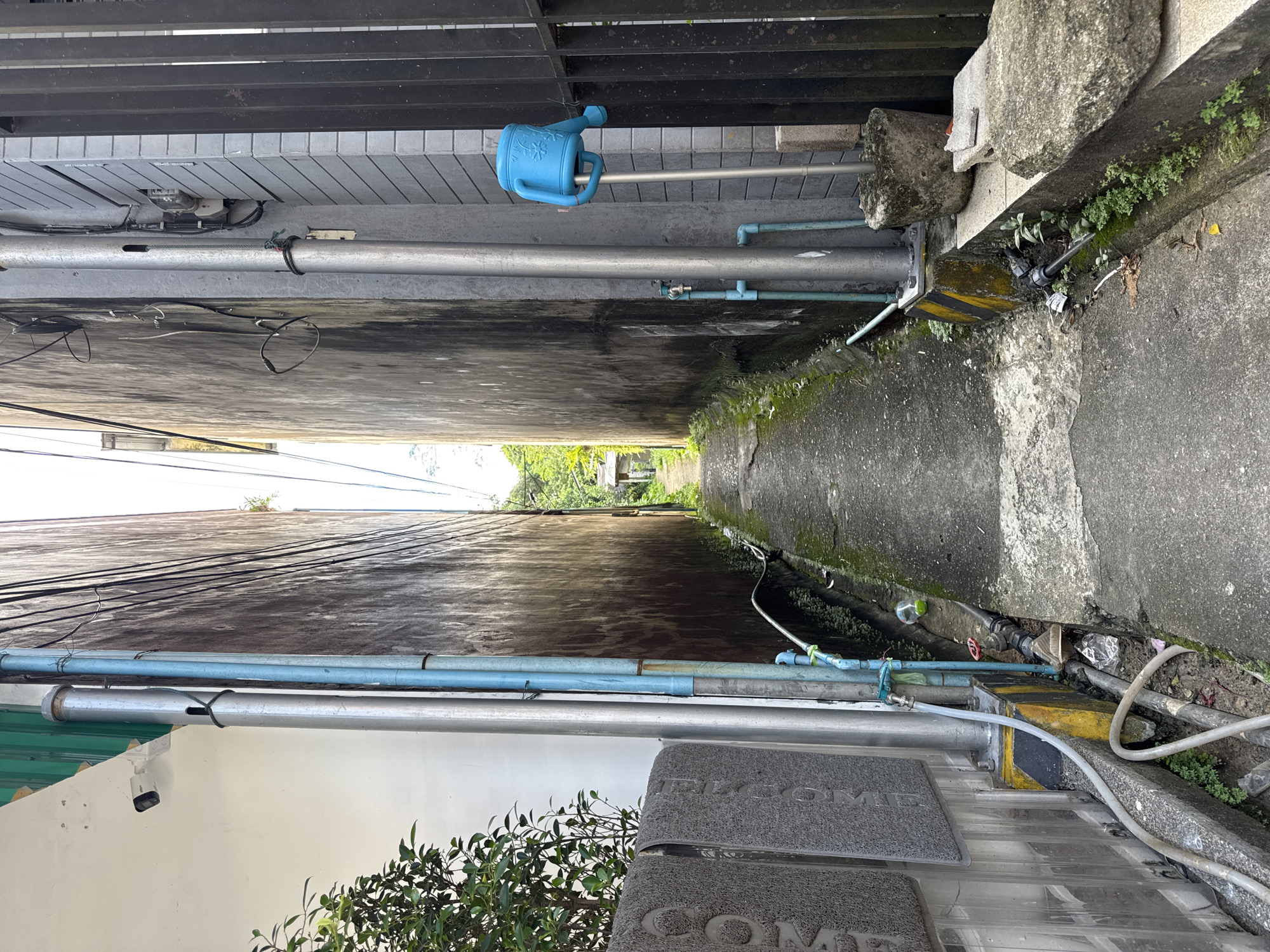 The image depicts a narrow alleyway in Ranong, Thailand, flanked by two tall, weathered concrete buildings. The alley floor is paved with asphalt and shows signs of wear, with patches of moss and weeds growing in the cracks and along the edges. Various pipes, some blue and others metallic, run along the walls, indicating utilities and water systems. On the left side of the alley, near the foreground, are two dark grey doormats with embossed patterns. A lush green plant with small leaves grows beside them.
On the right side of the alley, a light blue watering can is attached to a metal pole, positioned next to a black metal fence. The buildings themselves show signs of age and exposure to the elements, with discoloration and streaks on their surfaces. In the distance, the alley opens up to a brighter area, suggesting it leads to a more open space, possibly with vegetation and glimpses of small structures. The overall atmosphere is one of a utilitarian and somewhat neglected urban environment, common in many parts of Southeast Asia. There are no people visible in the image.