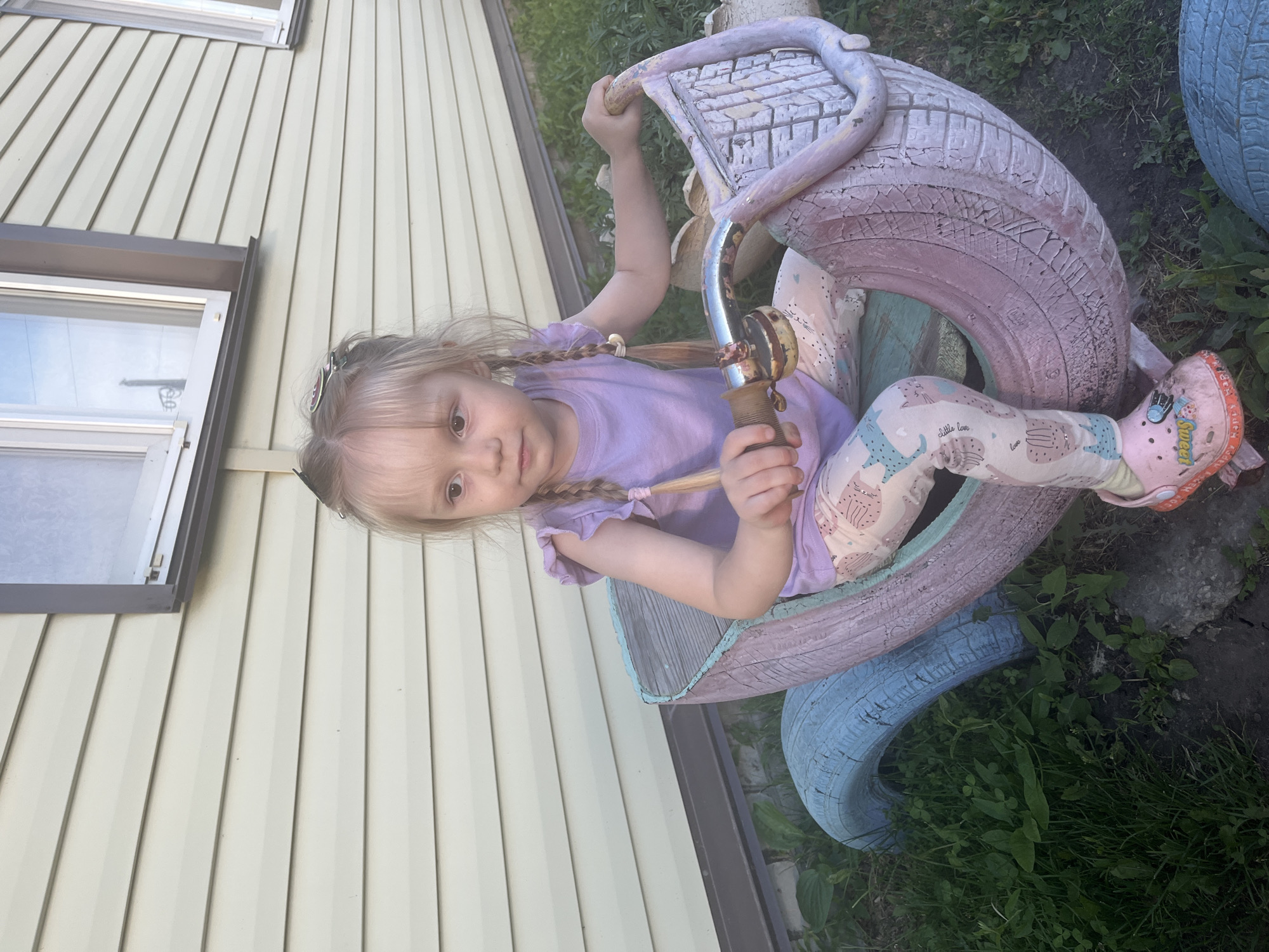 A young Caucasian girl with blonde hair in two braids is sitting on a toy vehicle made from painted tires. She is wearing a light purple t-shirt and patterned leggings. Her right hand is on the handlebars of the toy, which is decorated with a light blue and pink color scheme. The toy is situated on a grassy area with some visible rocks and a concrete border. In the background, a light-colored, horizontal-sided building with windows is visible. The image appears to be taken outdoors in daylight.